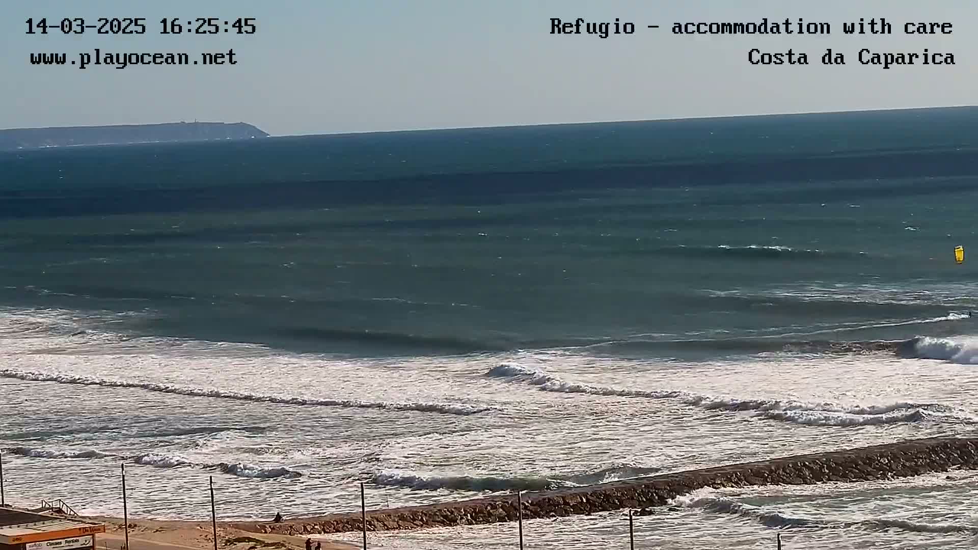 A long shot of ocean waves breaking on a shore under a clear sky, with a distant landmass visible on the horizon.