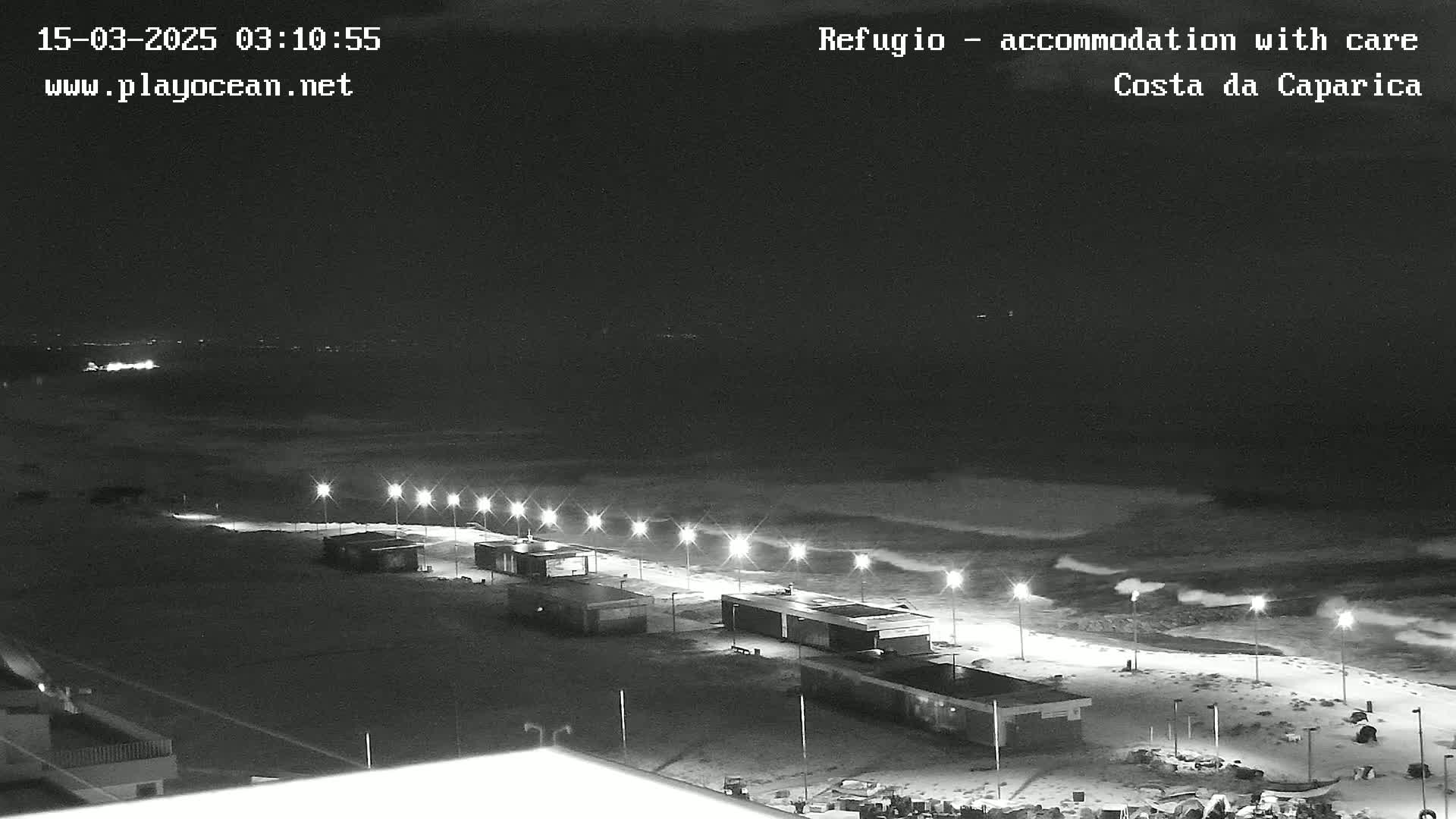 A nighttime beach scene with a row of lights illuminating a mostly empty beachfront area, and dark, wavy ocean under a clear, dark sky.