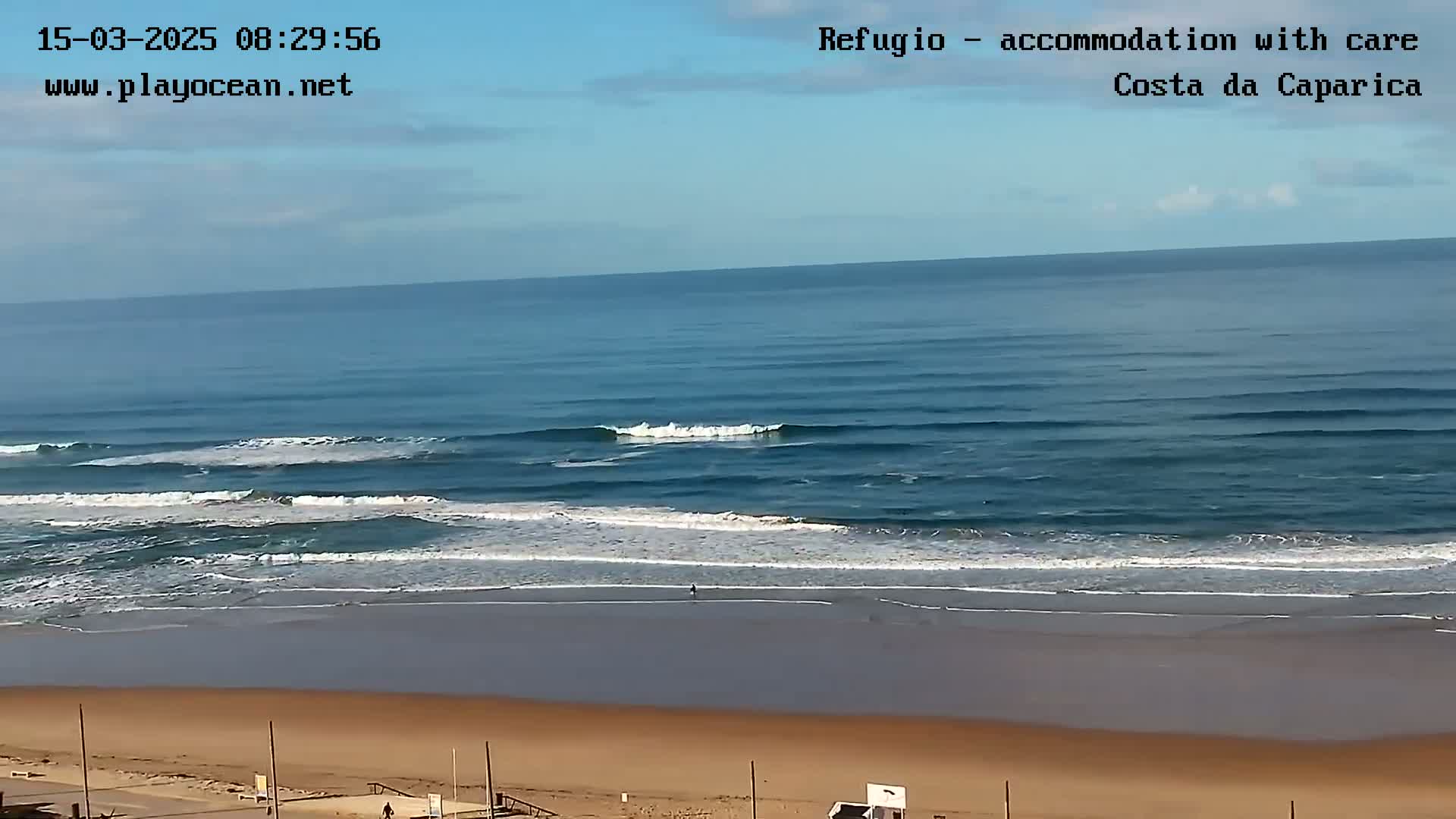 A mostly sunny day shows a wide sandy beach and ocean with small waves breaking near the shore.