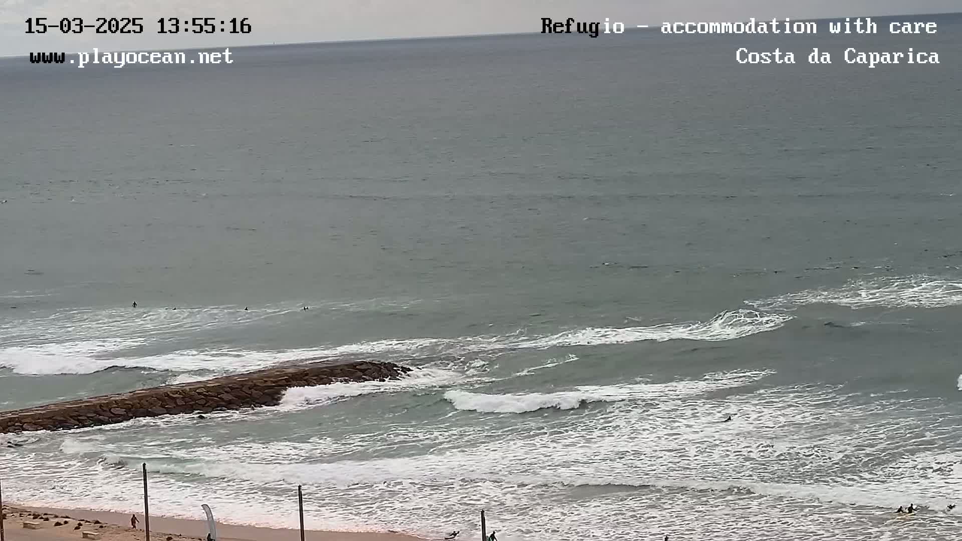 A high-angle, long shot shows a rocky jetty jutting out into a gray ocean with moderate waves under an overcast sky, and several people are visible surfing in the water and walking along the beach.