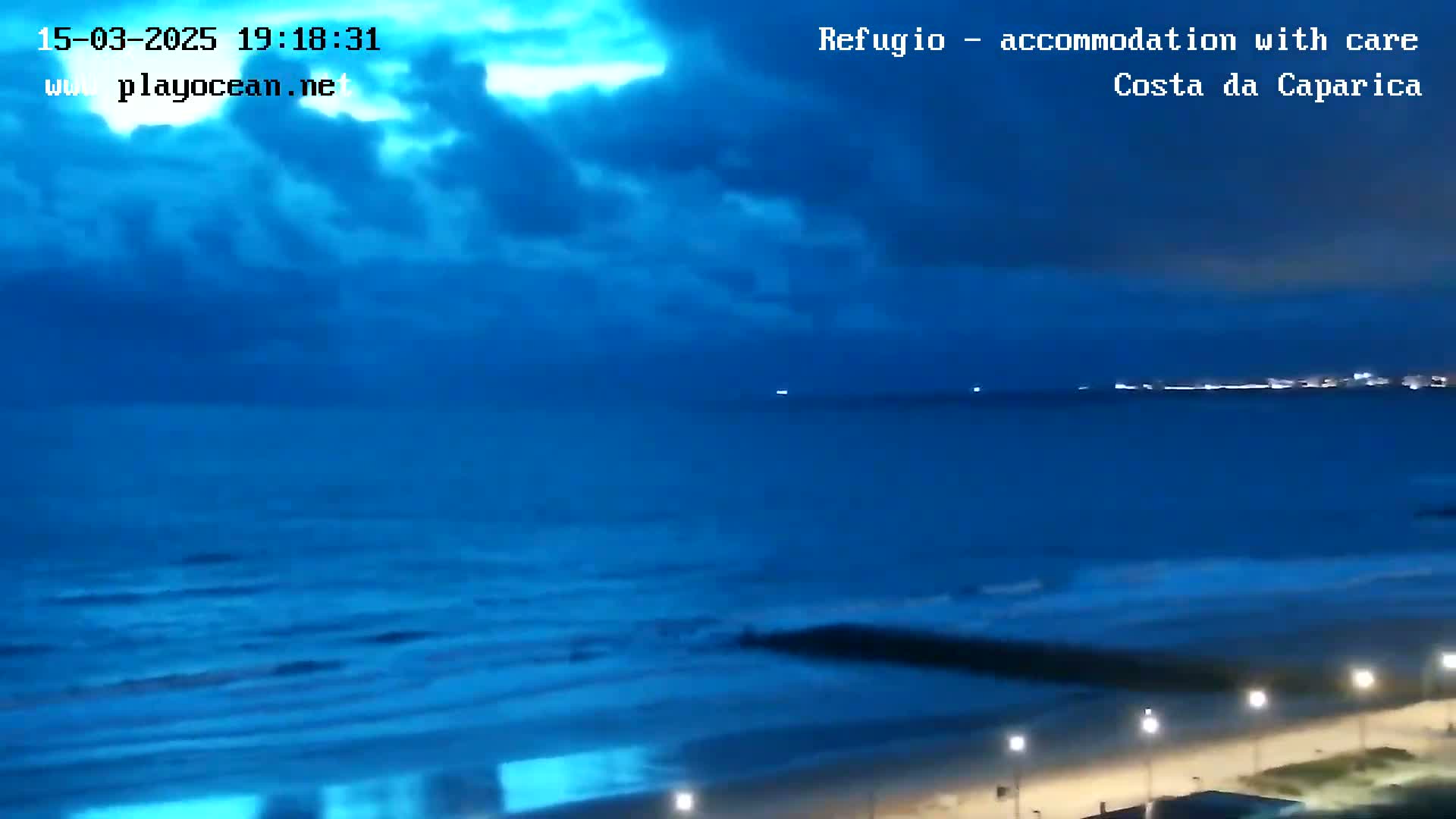 A dark blue ocean with small waves rolls onto a beach at night under a mostly cloudy sky.