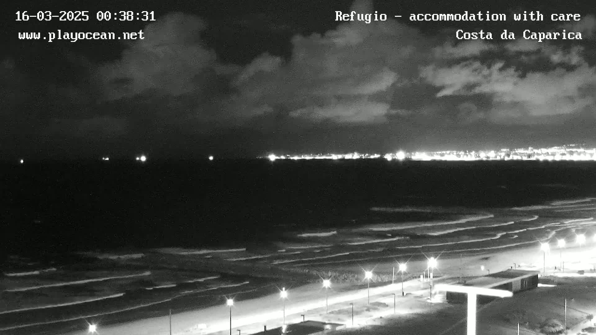 A nighttime view of a beach with gently rolling waves under a partly cloudy sky, showing a city's lights in the distance.