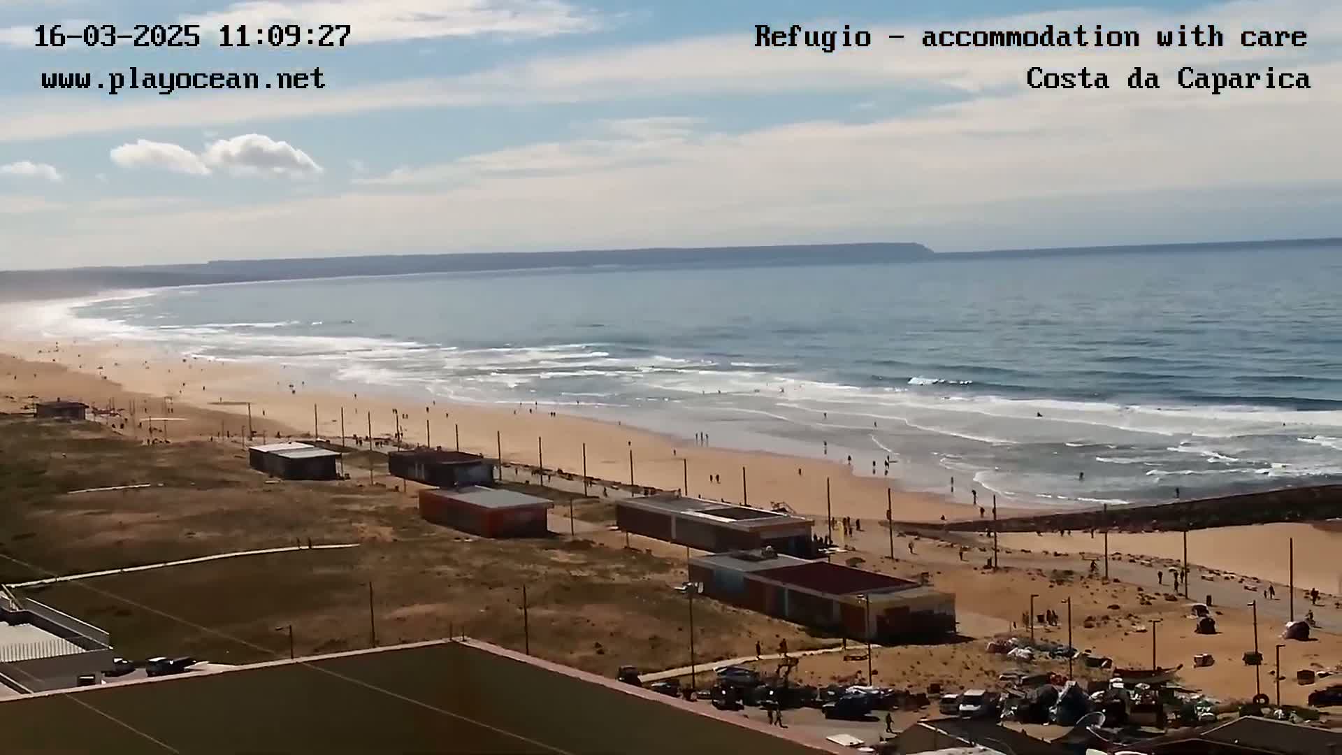 A mostly sunny day at a beach with small waves shows a wide sandy shore lined with small buildings and people, extending to a calm ocean meeting a distant headland under a partly cloudy sky.