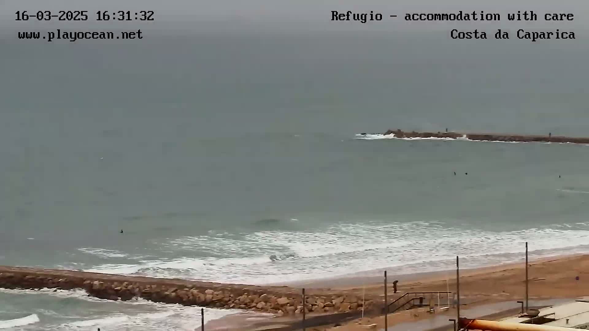 A mostly overcast day shows a sandy beach with moderate waves breaking against a low stone wall, a boat in the distance, and a few people near the water's edge.