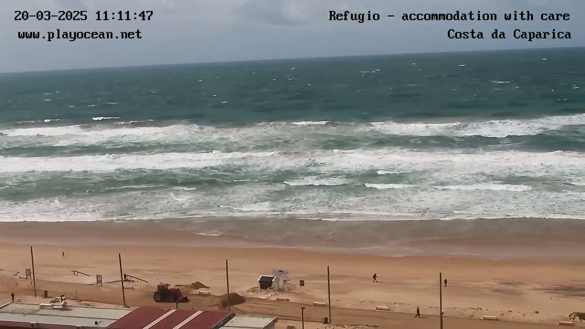 A sandy beach with a few people walking, a small tractor working near a lifeguard stand, and a somewhat choppy ocean with waves breaking near the shore under an overcast sky.