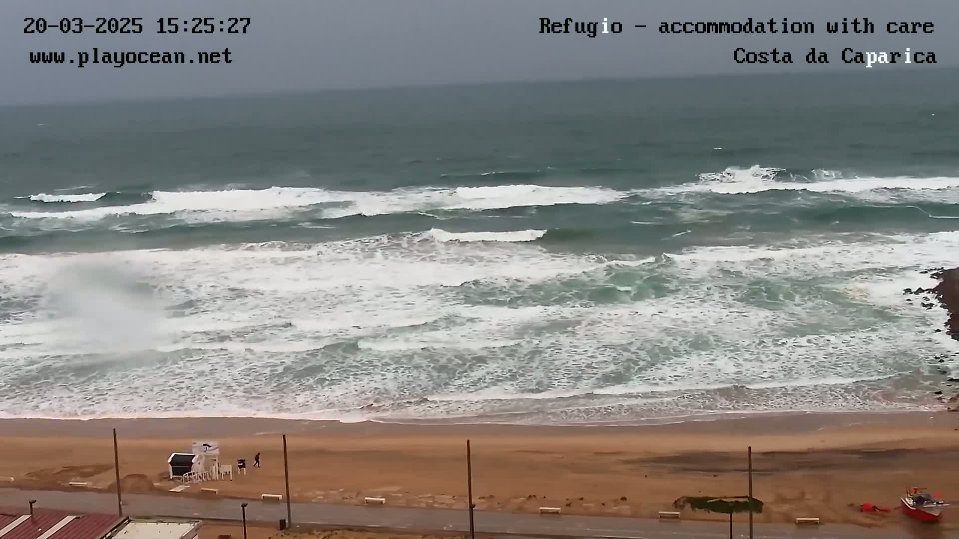 A sandy beach with a lifeguard stand and a few people is seen under an overcast sky, with moderately sized waves crashing on the shore.