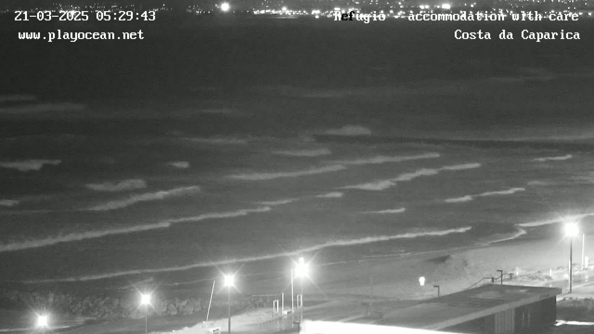 At night, gently rolling waves lap a dark beach under a clear sky, illuminated by several streetlights along a shoreline promenade.