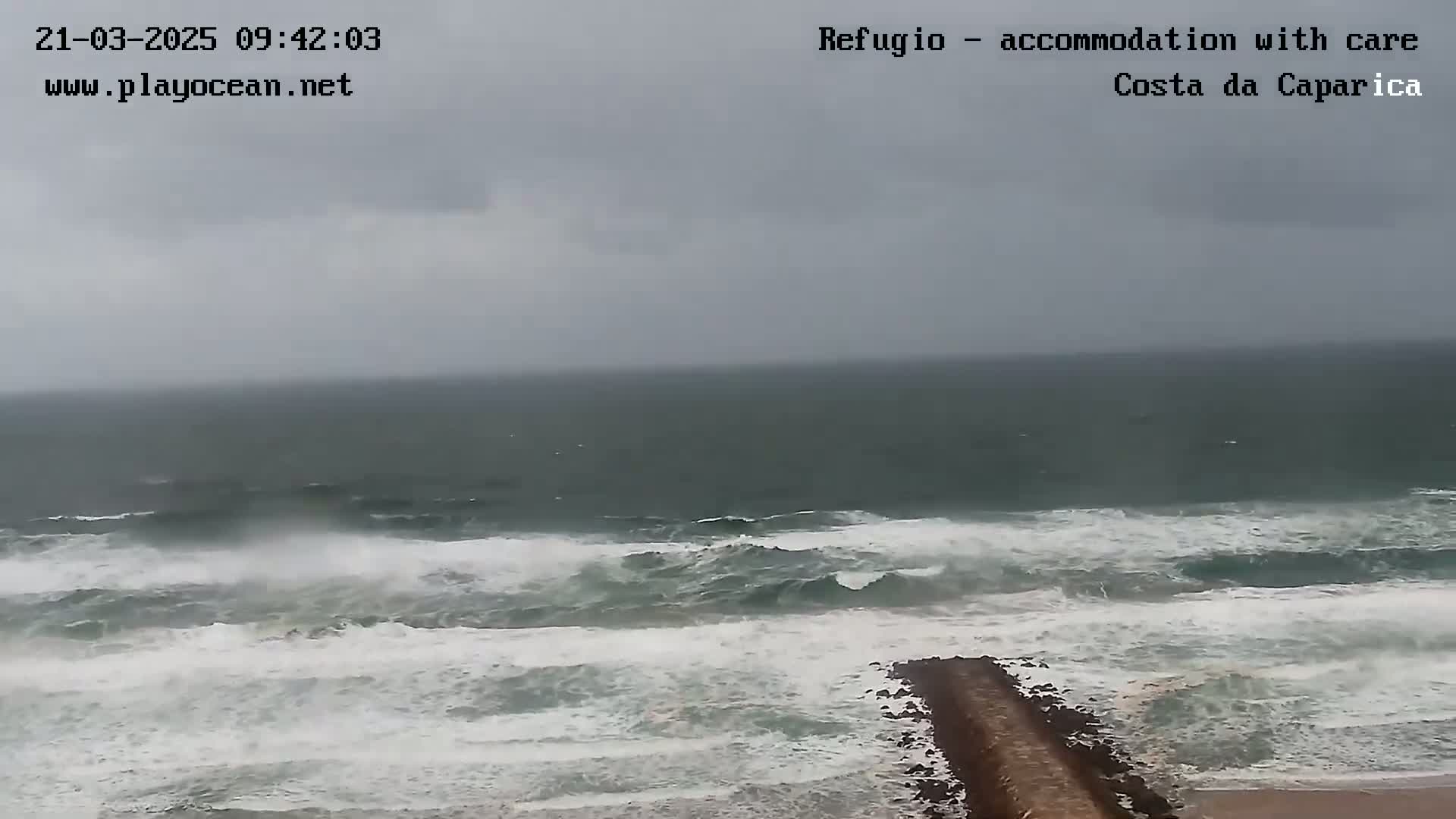 A long, dark-grey ocean with large, choppy waves crashes against a low-lying stone jetty on a cloudy day.