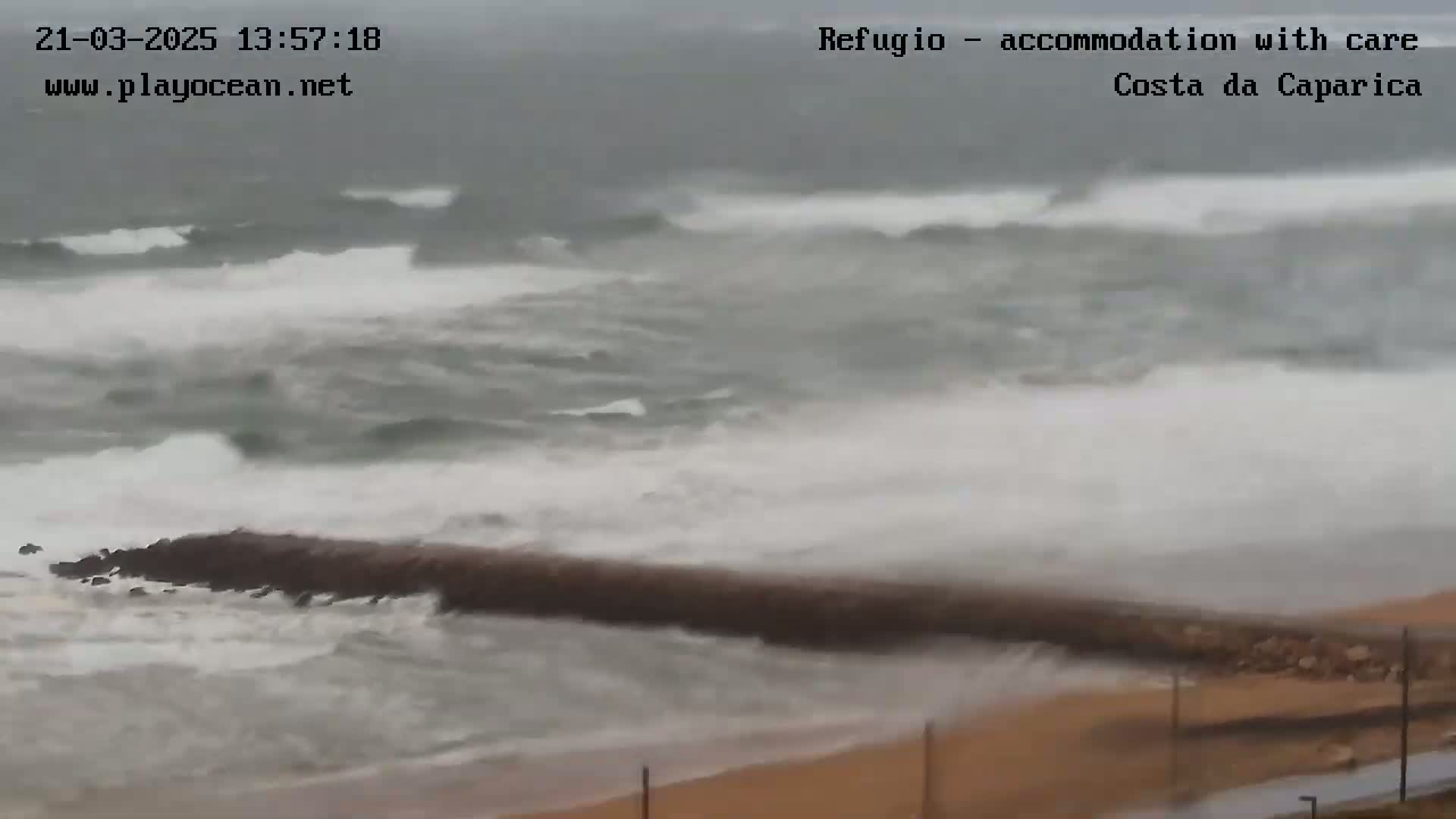 Large waves crash against a breakwater on a sandy beach during stormy weather.