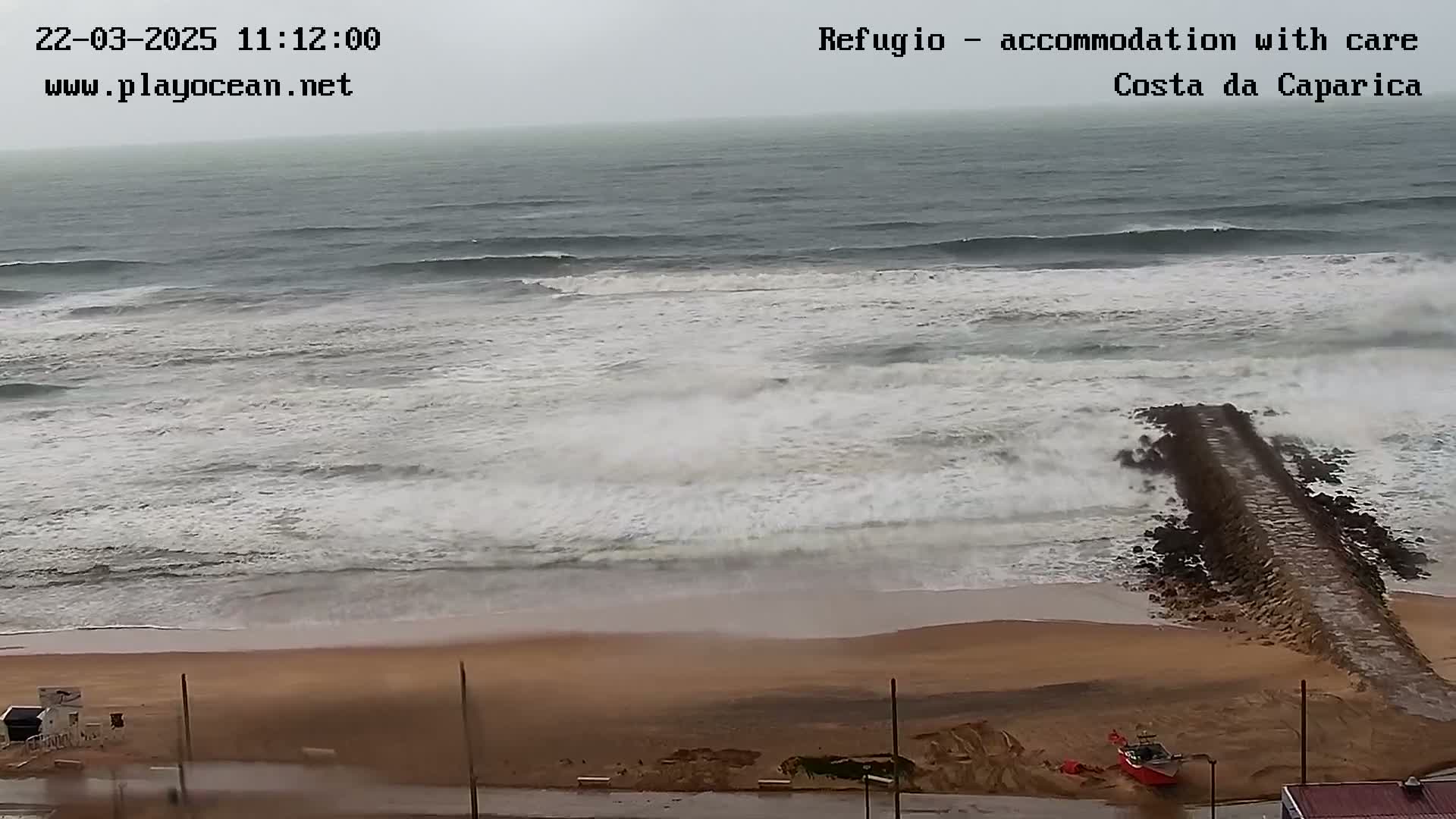 A sandy beach with a rocky jetty extending into a rough, gray sea under overcast skies.
