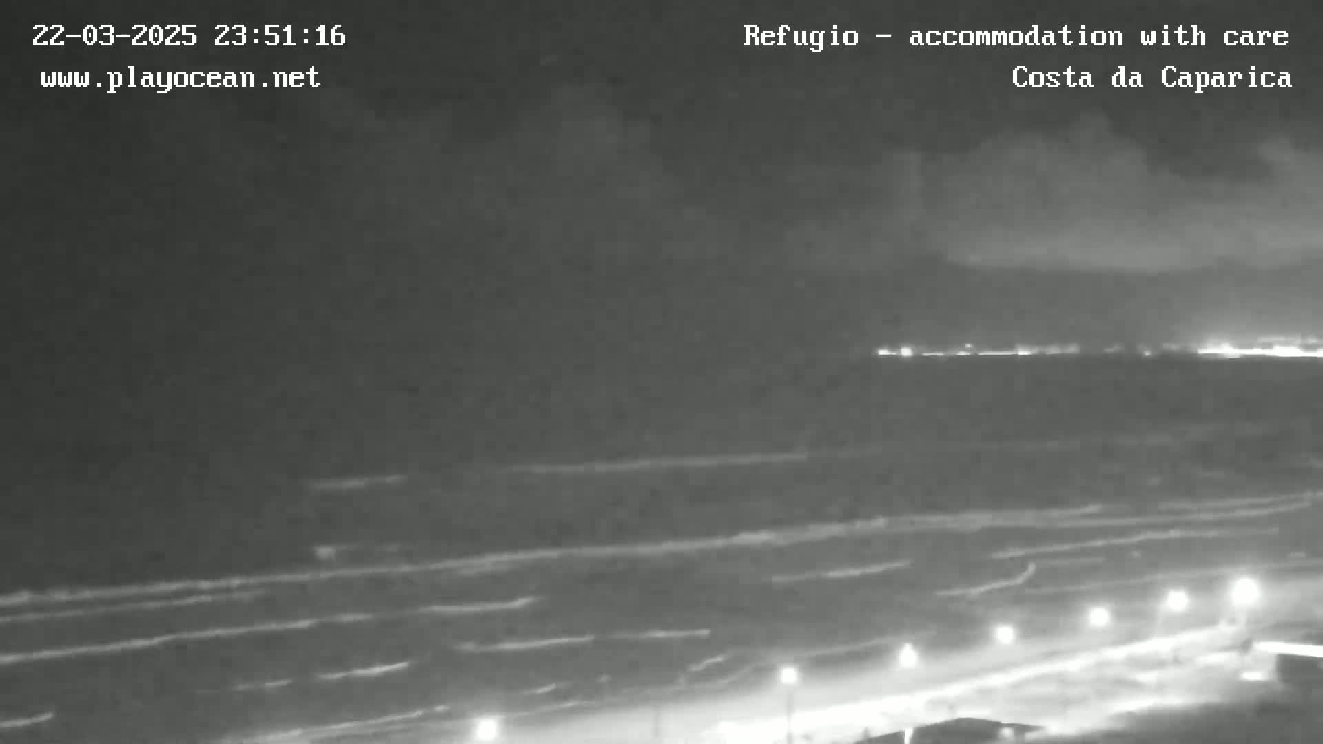 A nighttime, overcast view of a beach with gently rolling waves and lights visible along the shore.