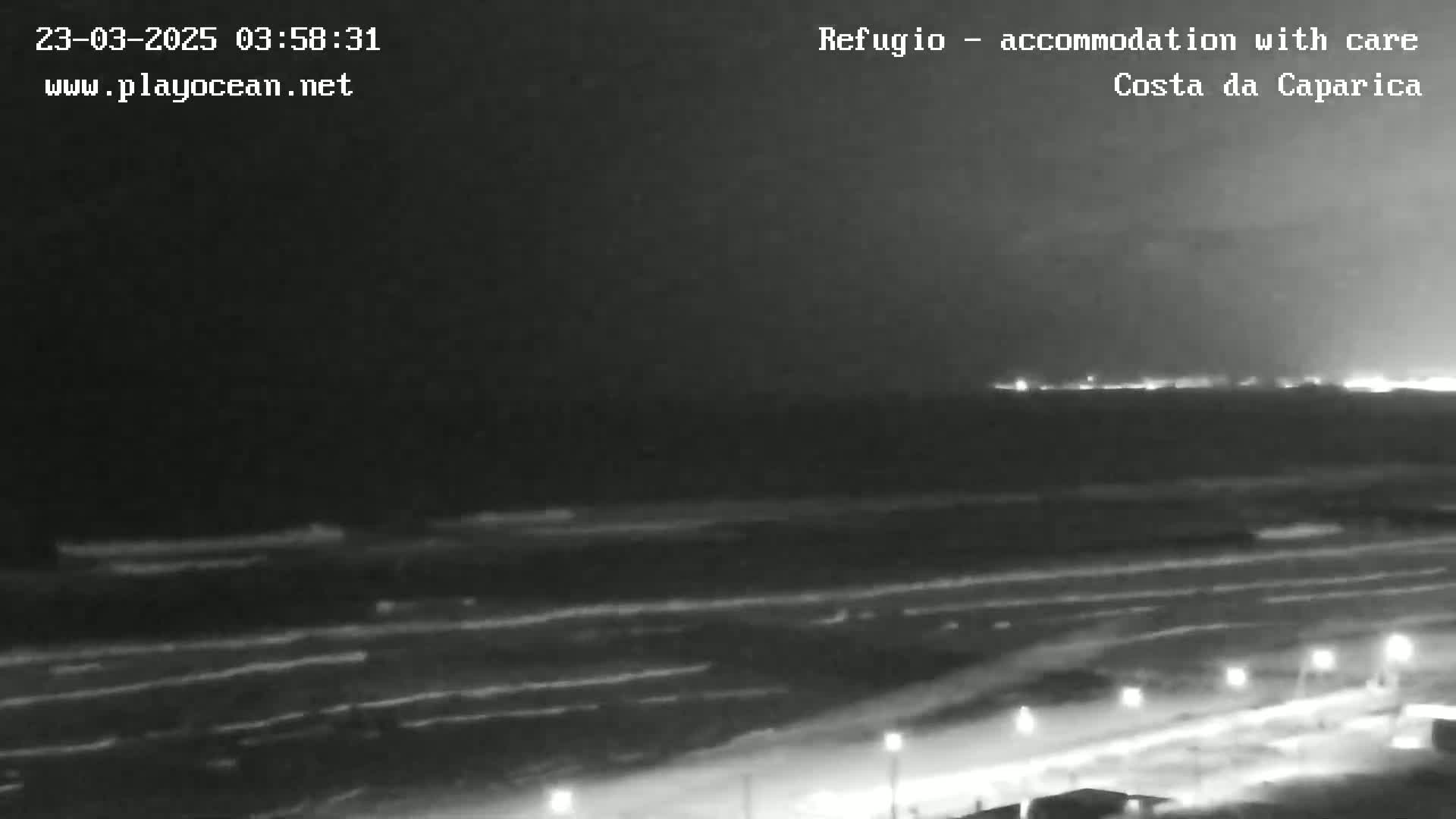A nighttime view of a dark beach with gently rolling waves under a cloudy sky, and lights visible in the distance along a coastline.