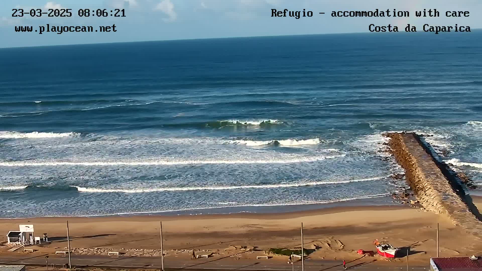 Ocean waves roll onto a sandy beach next to a long, low stone jetty under a partly cloudy sky.