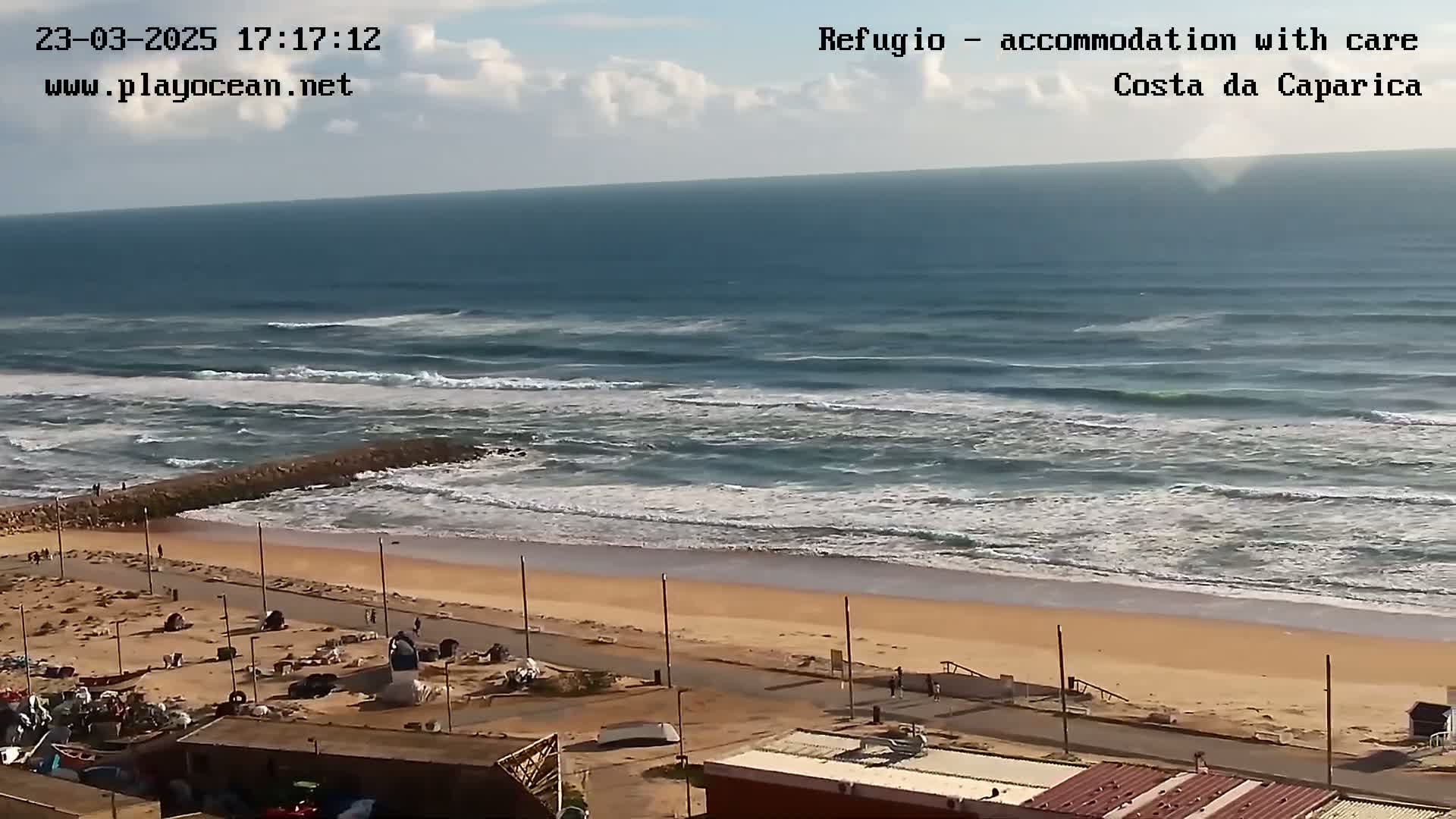 A sandy beach with a rocky jetty and moderate waves under a partly cloudy sky is viewed from a high vantage point, showing buildings and a roadway in the foreground.