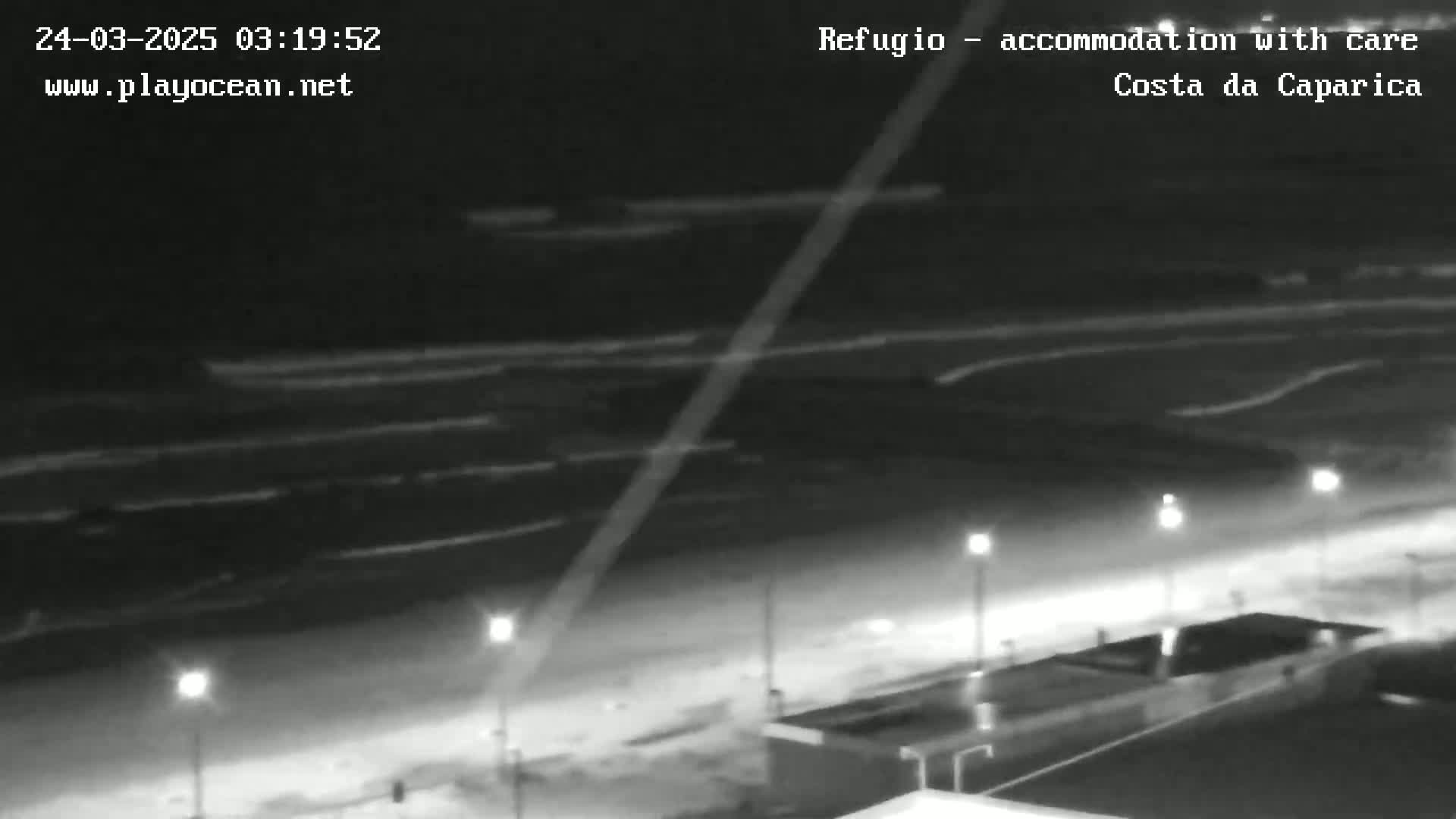 A nighttime, black and white view of a beach with gentle waves rolling onto the shore, illuminated by streetlights along a nearby walkway.