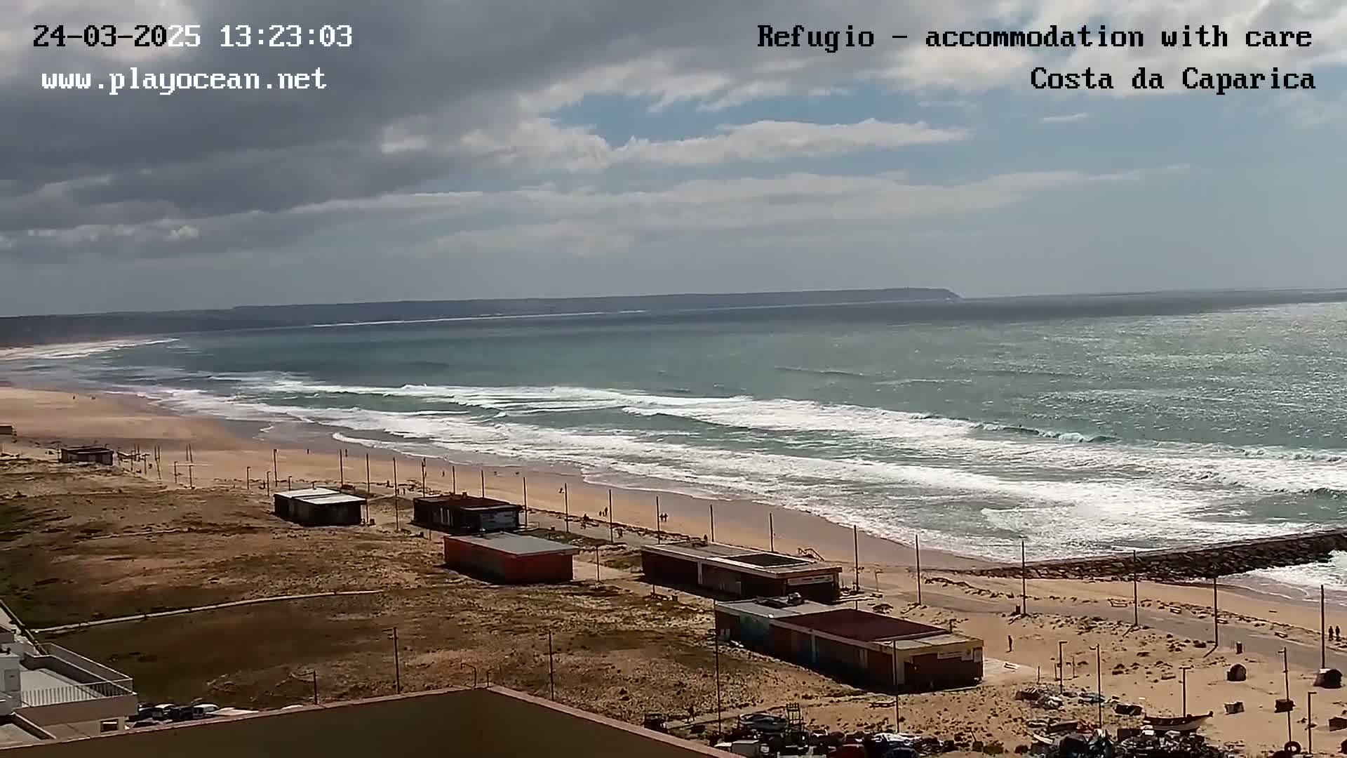 A wide shot shows a sandy beach with small buildings and a calm ocean under a partly cloudy sky.
