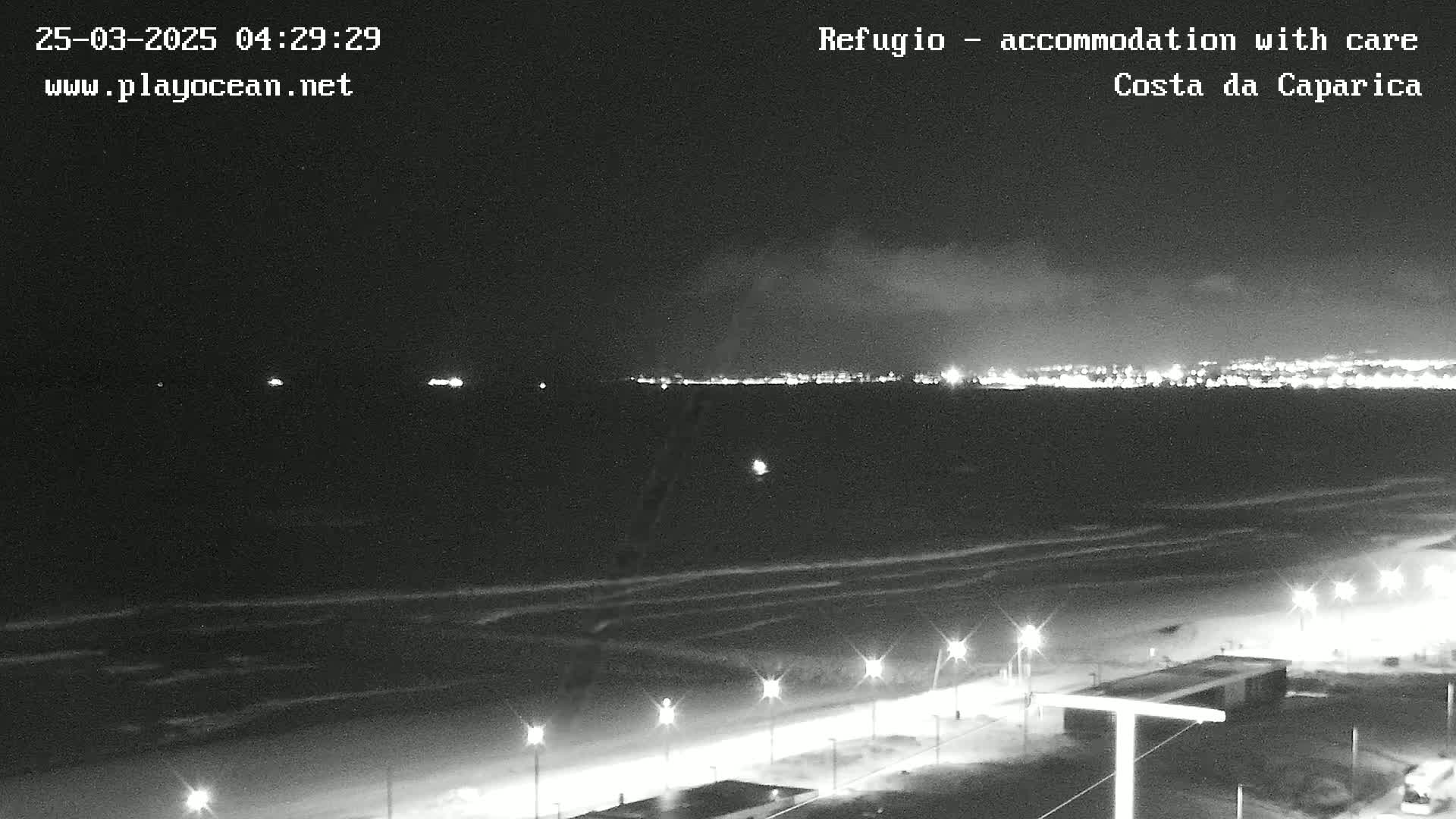 A nighttime, overcast view of a dark ocean beach with lights illuminating a shoreline path and a distant city skyline.