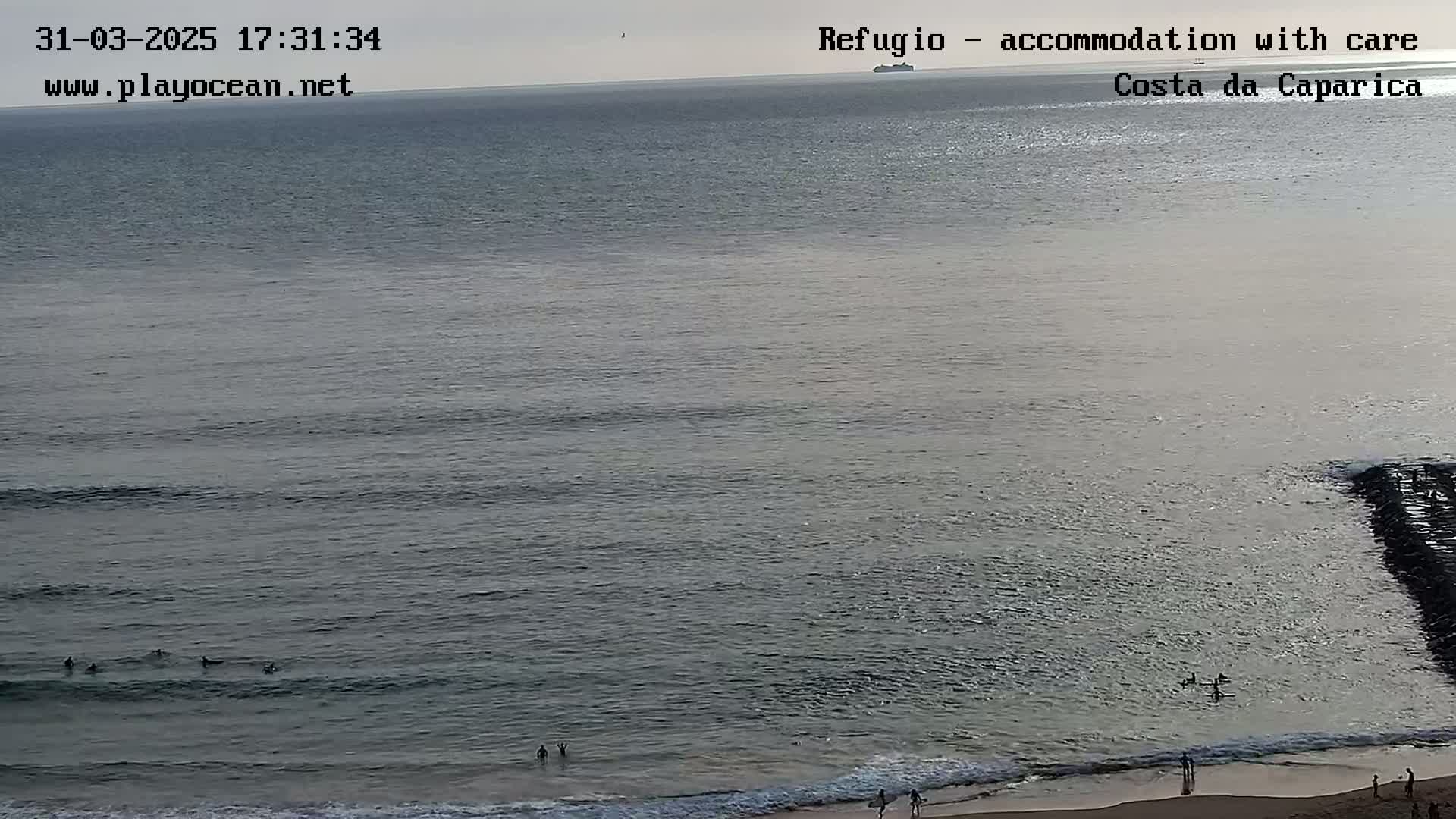 A calm ocean with several surfers in the water and people on a sandy beach under a cloudy sky.