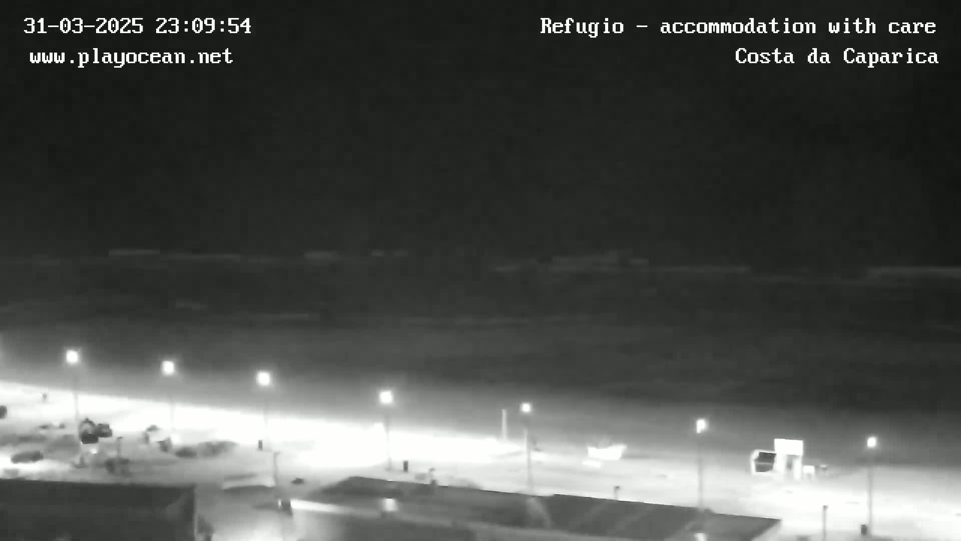 A nighttime view of a dark beach with calm water and sparsely lit beachfront buildings.