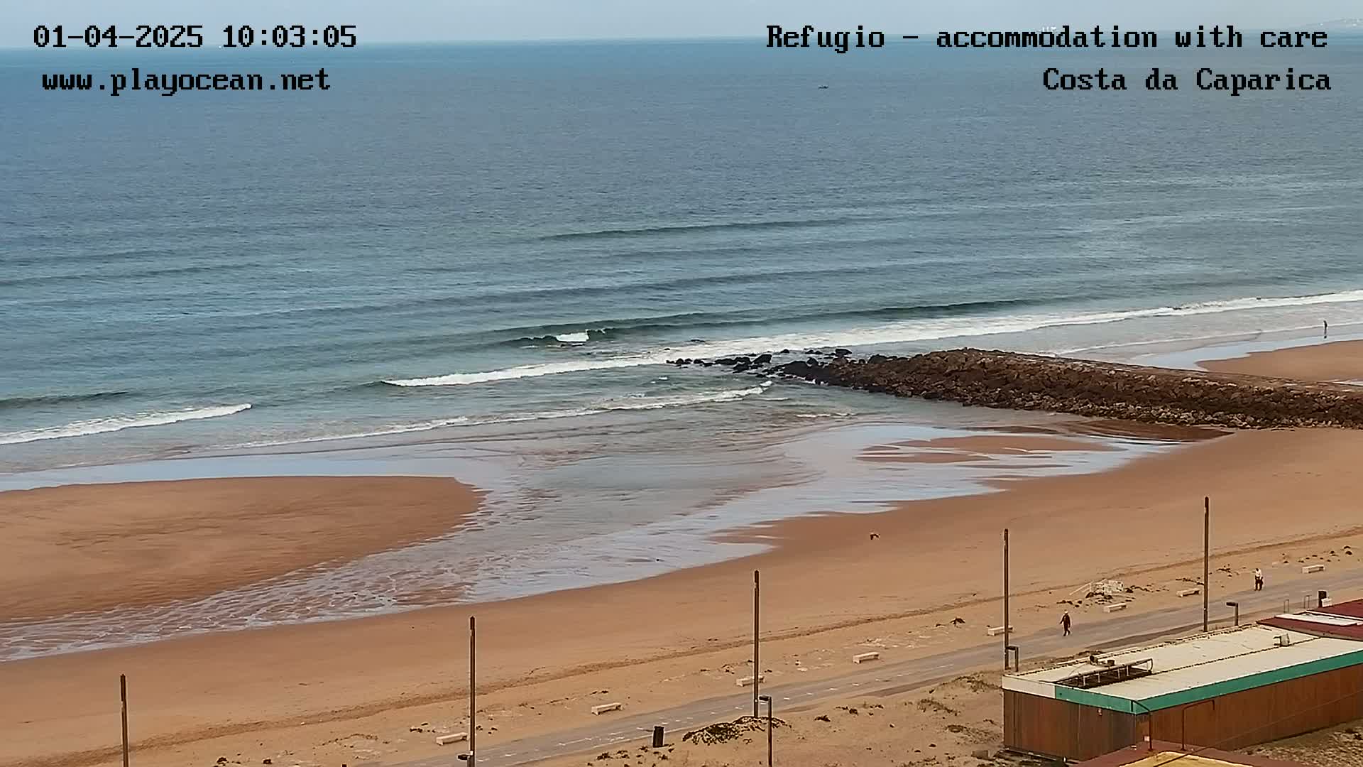 A sandy beach with small waves rolling in on an overcast day, a rock jetty partially separating the water from the shore, and a paved road running along the beachfront.