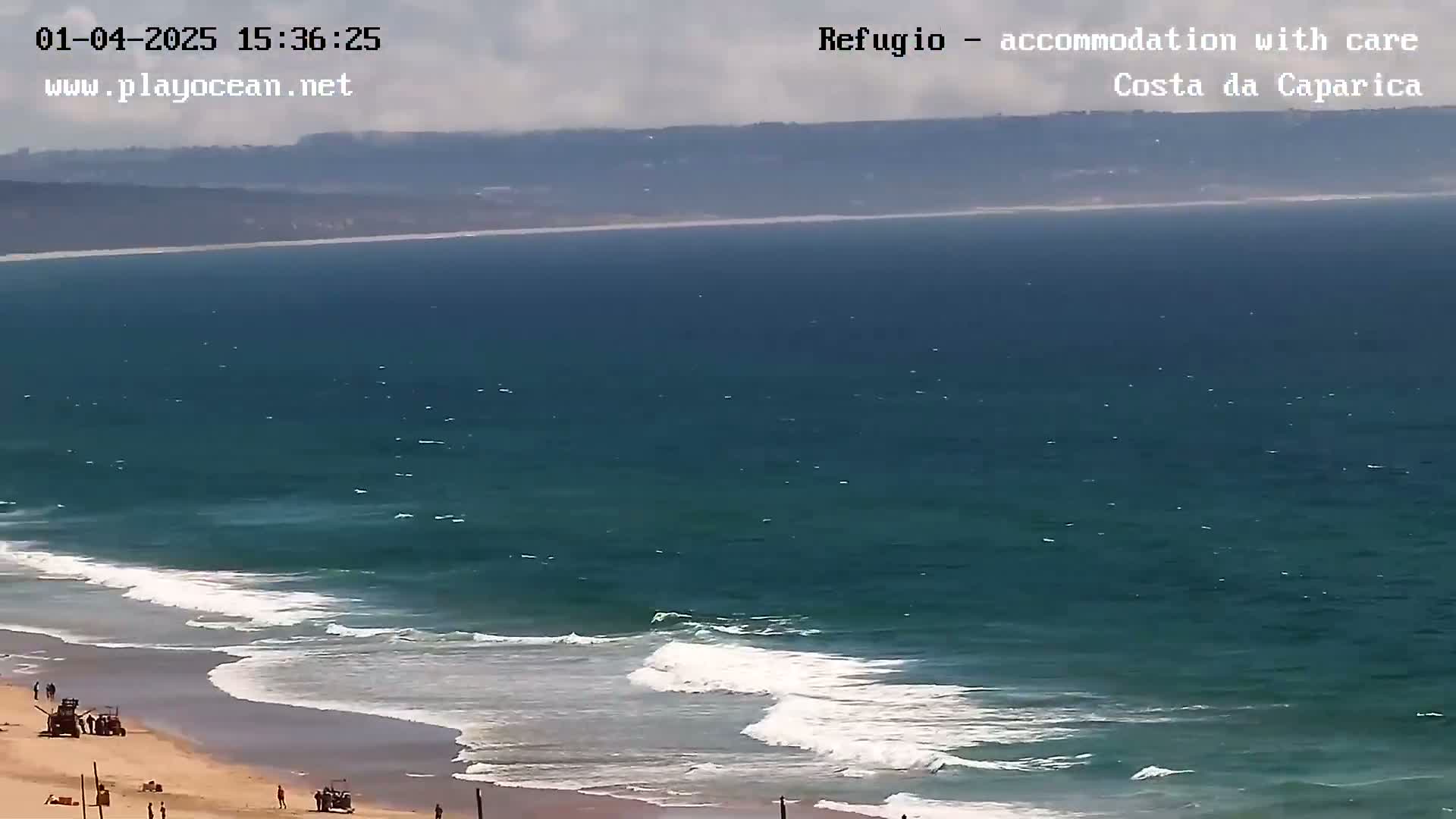 A sandy beach with a few people and vehicles is visible, with waves rolling onto the shore from a dark-teal ocean under a partly cloudy sky.