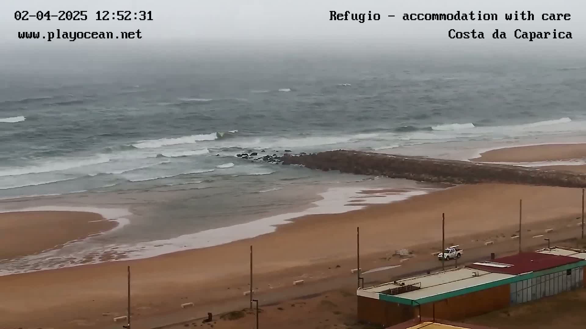 A sandy beach with a rocky breakwater is visible, with overcast skies and moderate waves rolling into shore.