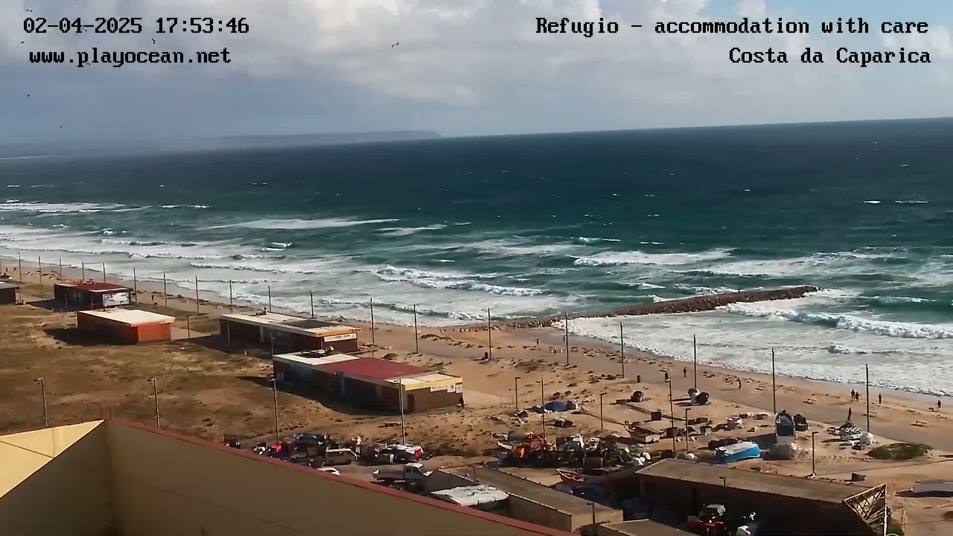 A beach with several small buildings and a parking lot is shown, with a choppy ocean and partly cloudy skies in the background.