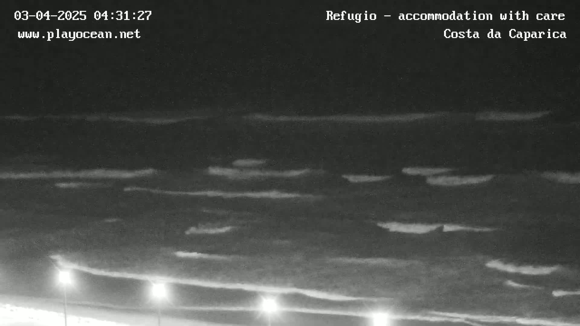 A nighttime, low-light image shows a dark ocean with faint, horizontal cloud formations above it, and several lights are visible along a dark shoreline at the bottom of the image.