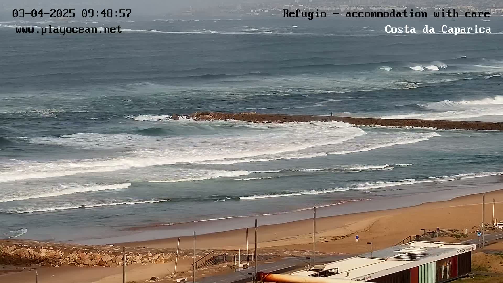 An overcast day shows a long sandy beach with moderate waves breaking over a low-lying rock wall that extends into the ocean.