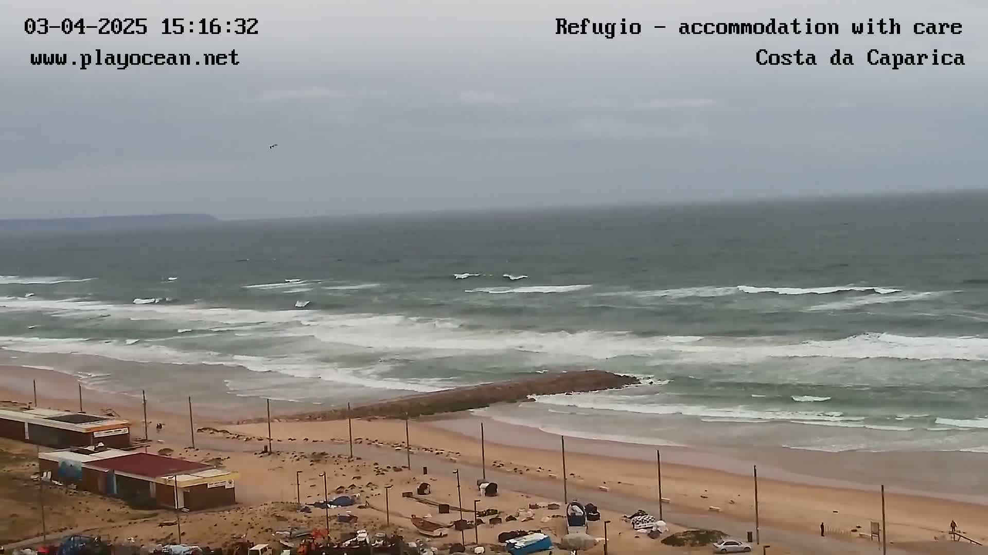 An overcast day shows a beach with moderate waves breaking near a shoreline with small buildings and some vehicles parked nearby.