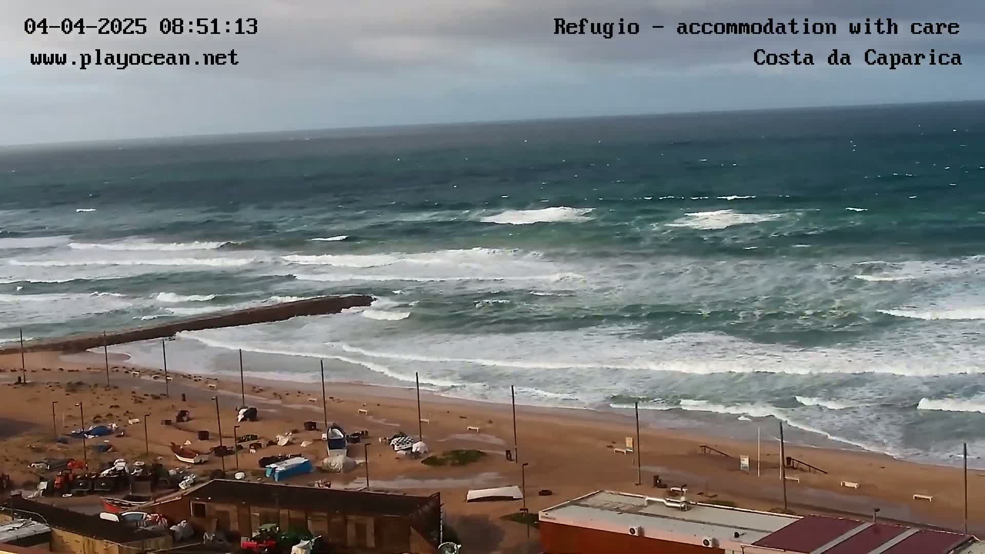 A beach with choppy waves crashing on the shore under an overcast sky, showing some debris on the sand and boats near the buildings.