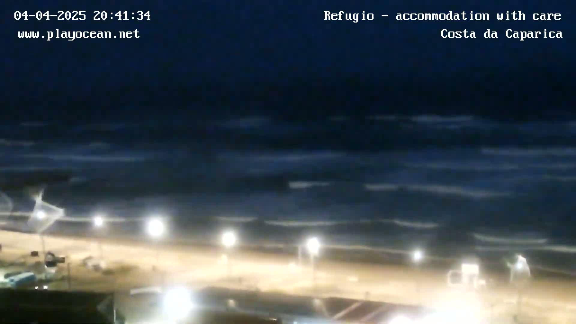 A nighttime view of a beach with dark, wavy ocean water under a mostly clear, dark sky, illuminated by several lights along the shoreline.