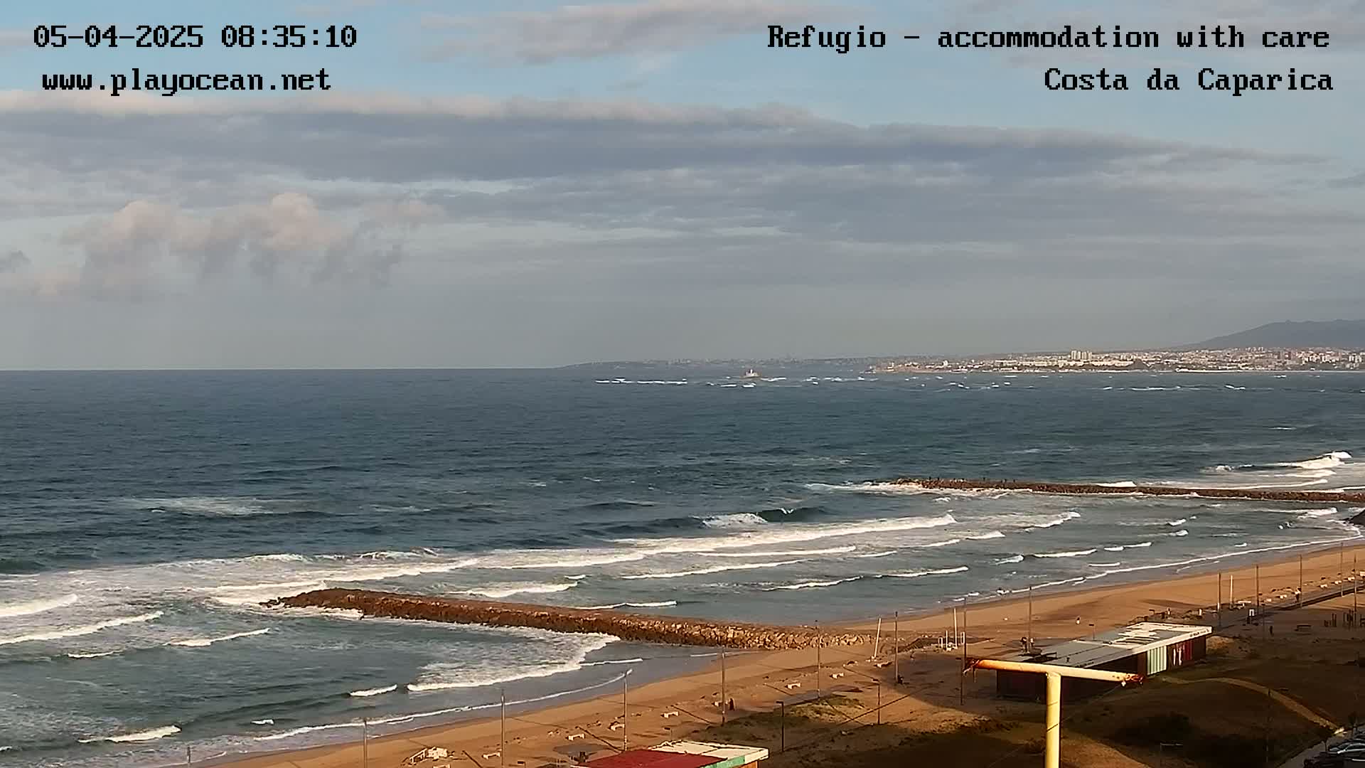A partly cloudy day shows a view of a beach with waves breaking against a rock jetty, and a town visible in the distance.