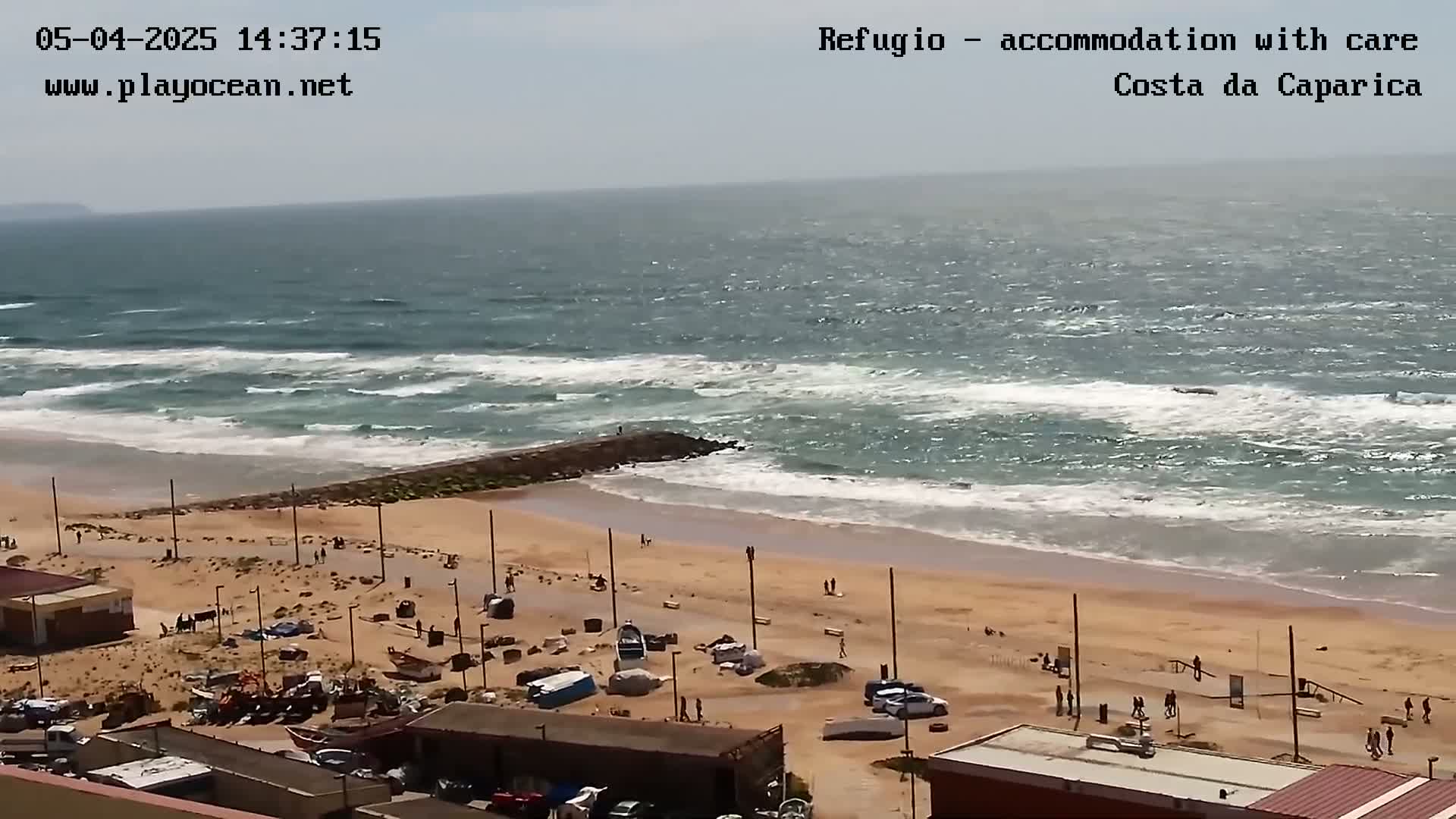 A sandy beach with a rocky jetty, moderate waves, and a few people are visible under a mostly clear sky.
