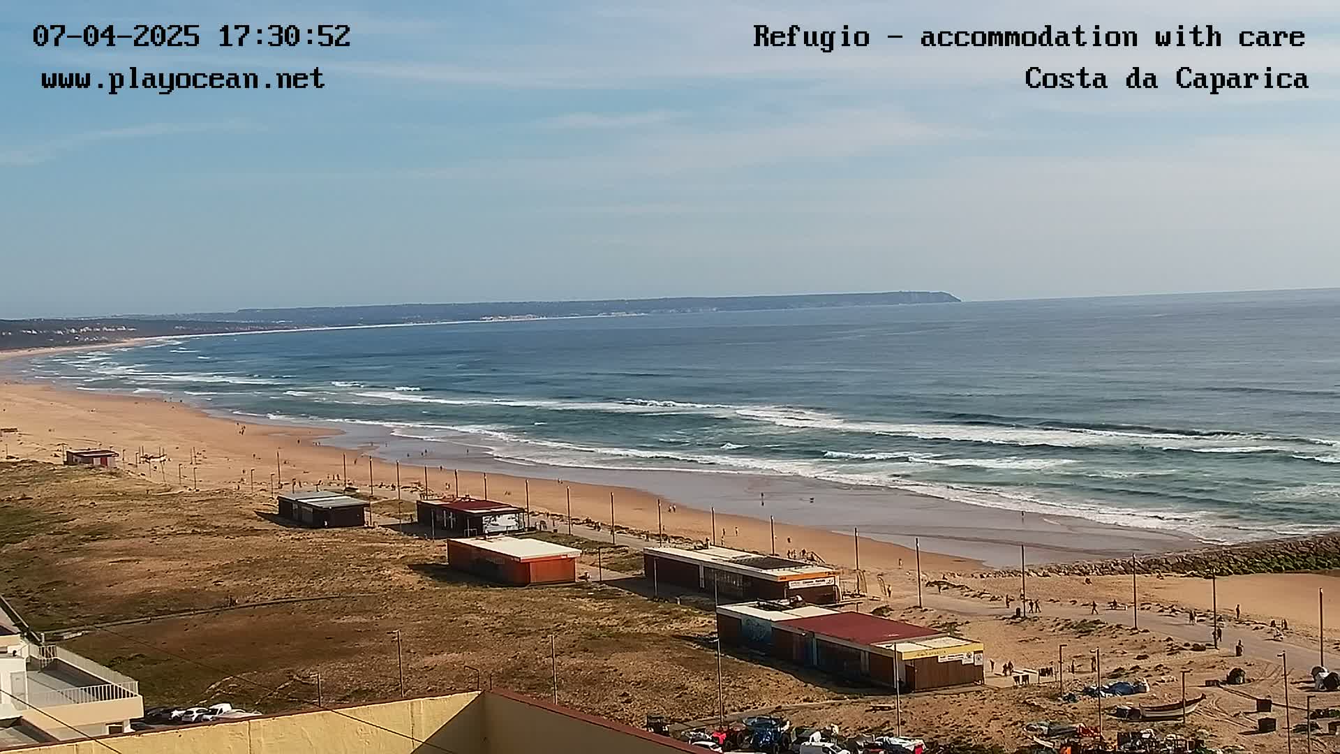 A long sandy beach with small buildings and people, rolling waves in a calm ocean under a clear blue sky.