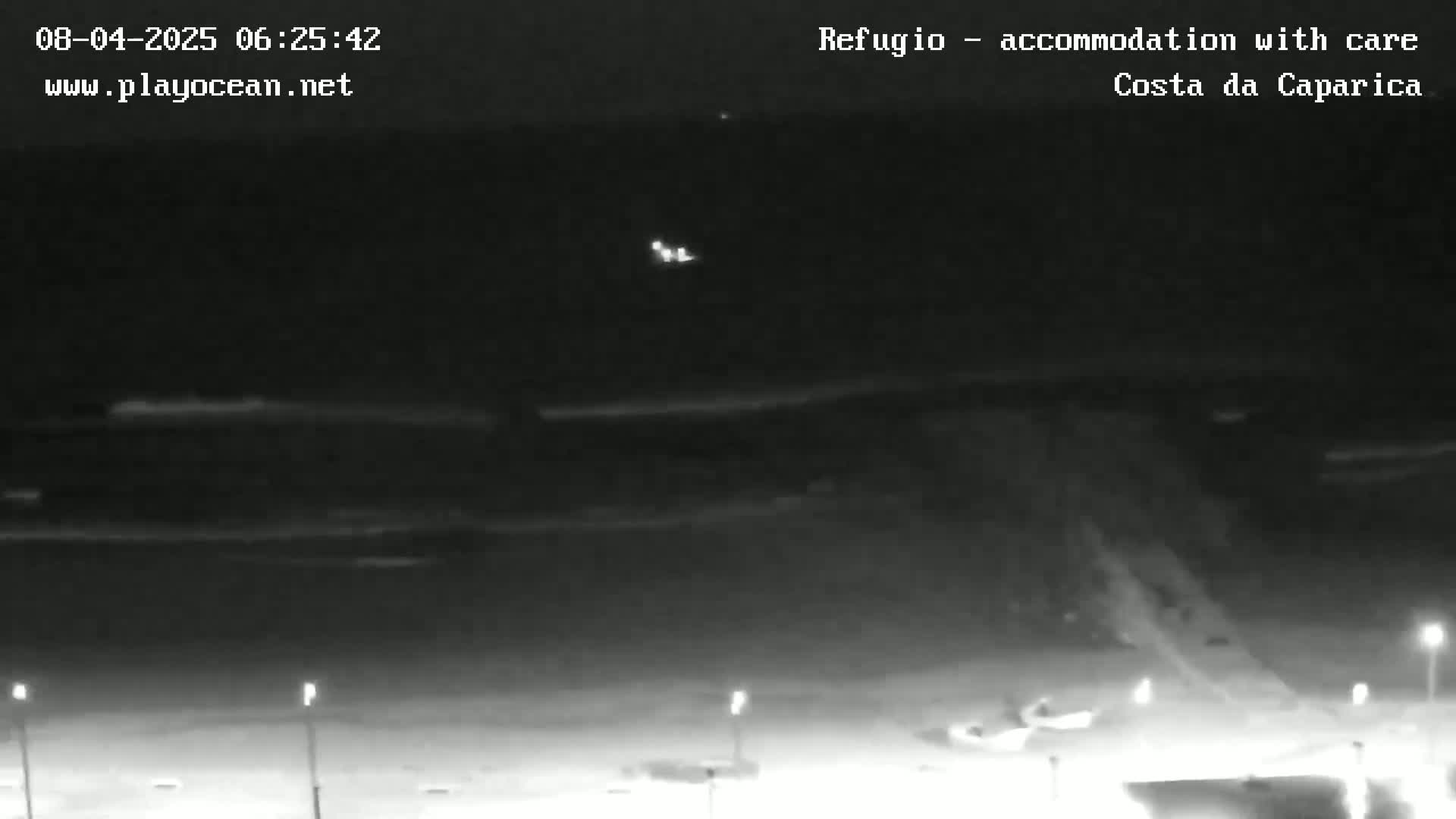 A nighttime, low-light view of a beach with dimly lit structures and a dark ocean with faint waves under a mostly clear sky.