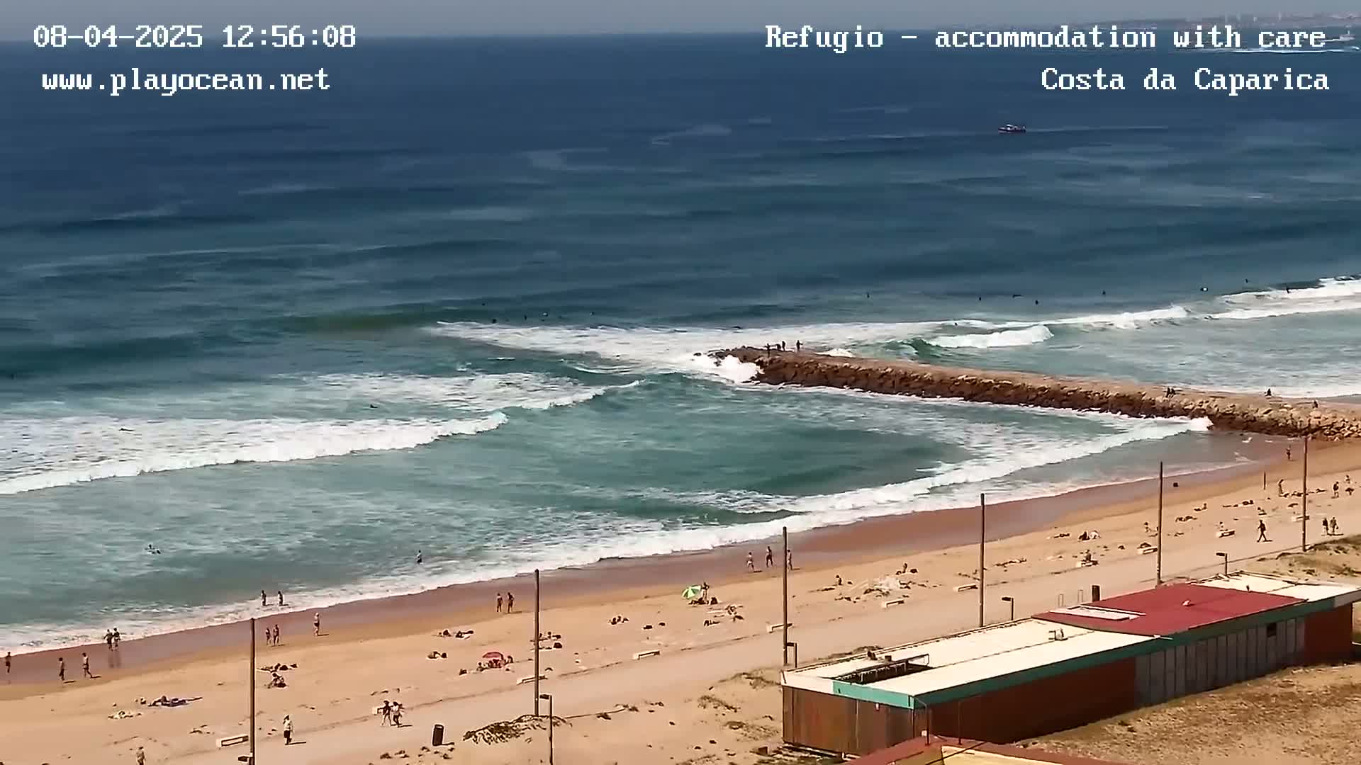 A sandy beach with people sunbathing and swimming is seen next to a rocky jetty where waves break, all under a clear sky.