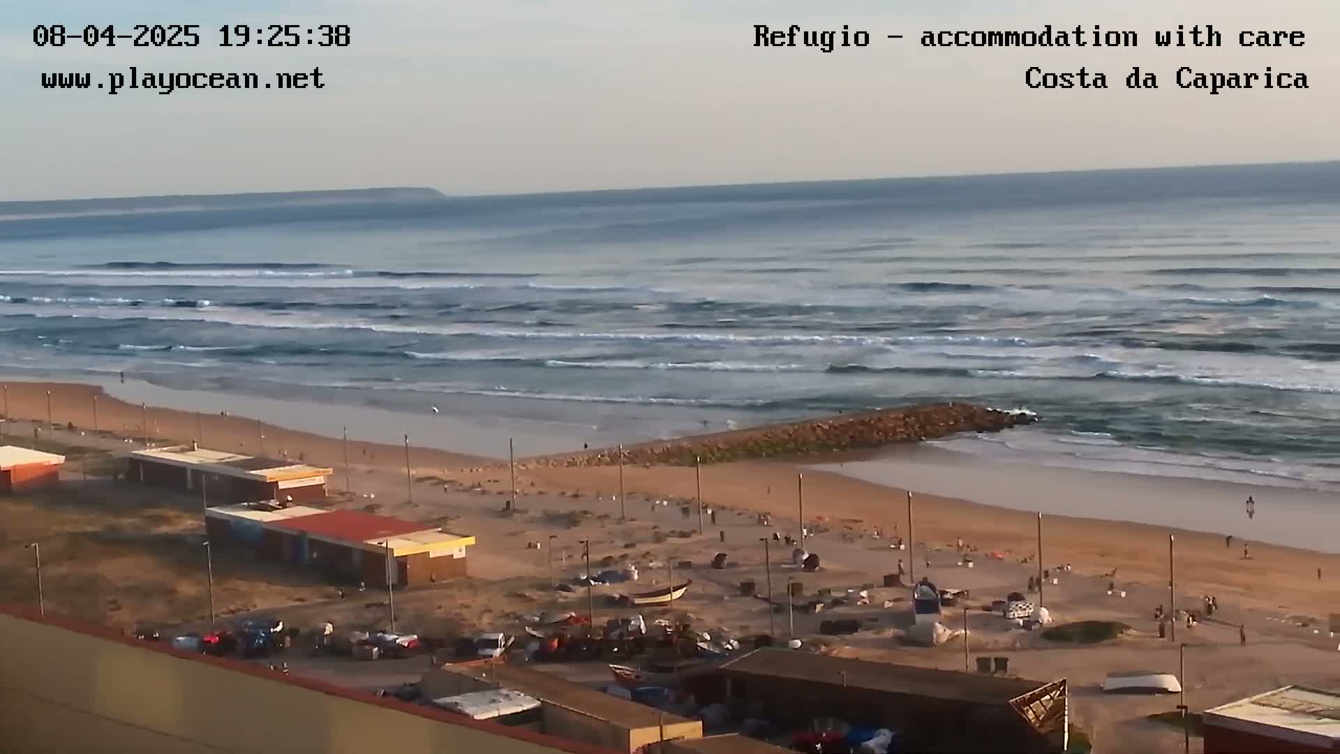 An ocean beach with moderate waves under a mostly clear sky shows people on the sand and near small buildings.