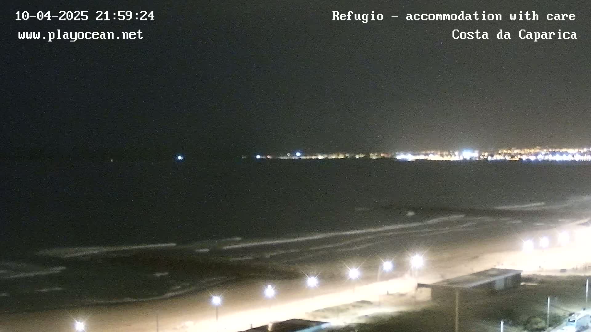 A nighttime view of a beach with gentle waves, illuminated by streetlights, and a distant city skyline visible across the water.