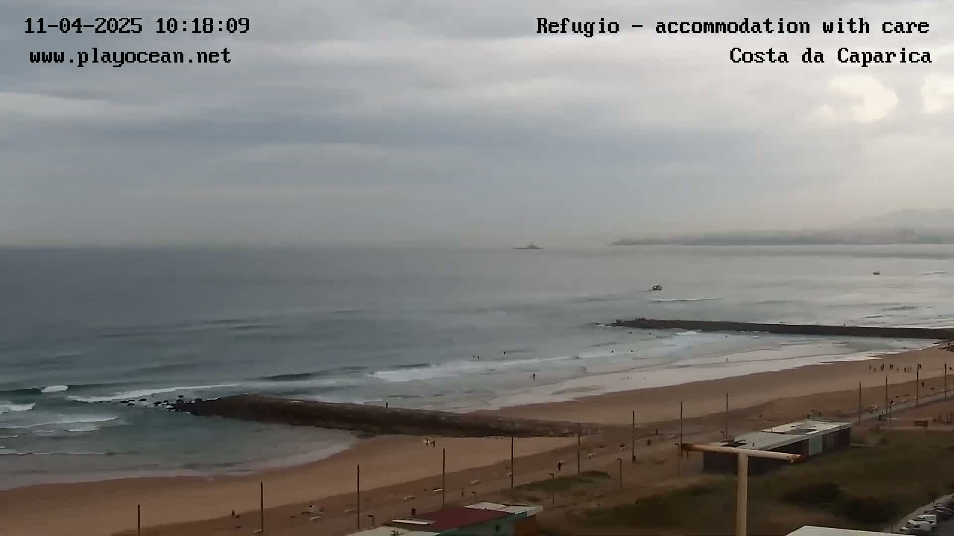 A sandy beach with small waves under an overcast sky is seen from an elevated vantage point, showing a rocky jetty extending into the ocean.