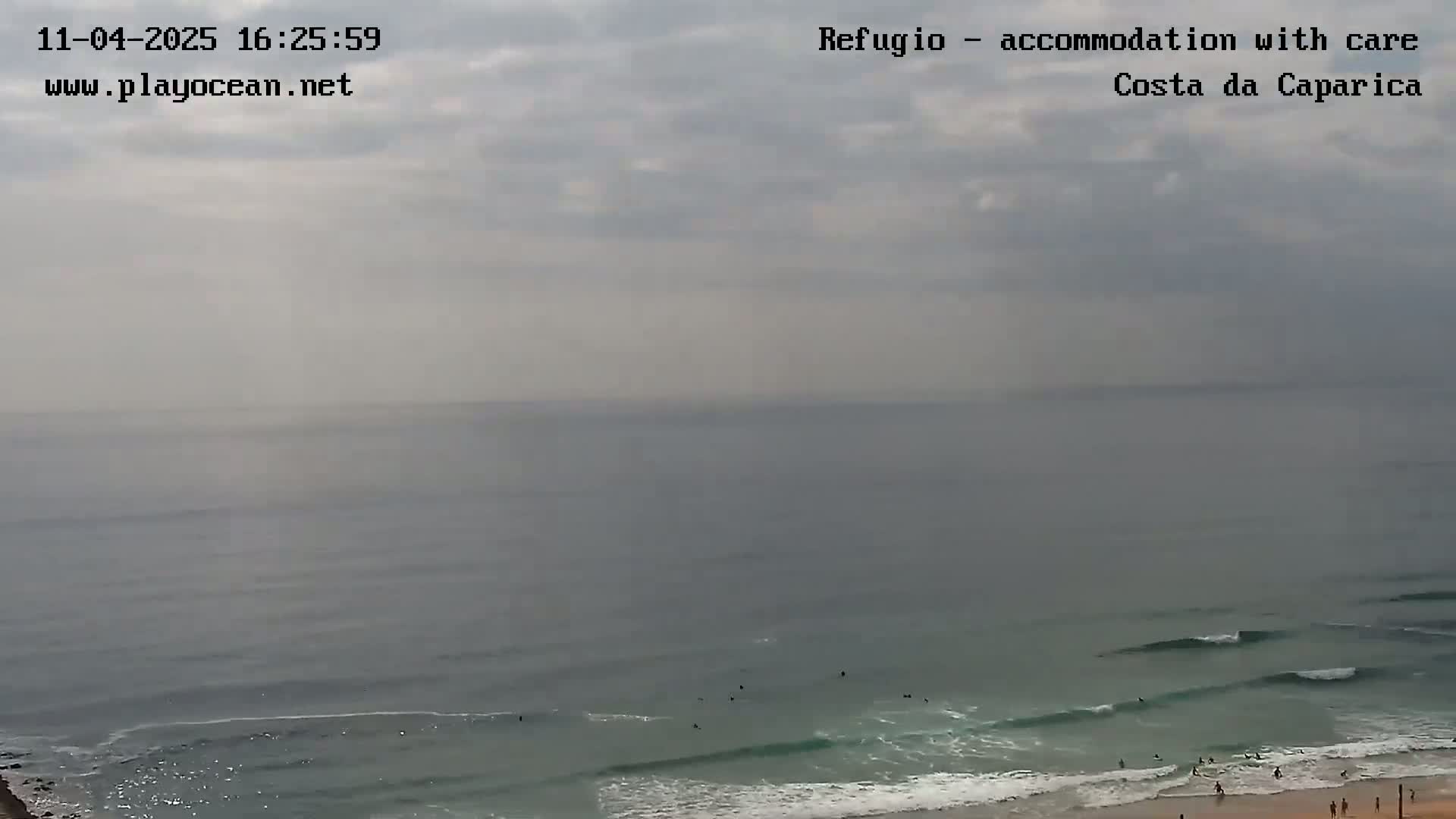 A mostly cloudy day shows a wide expanse of ocean with small waves and a few surfers near the shore.