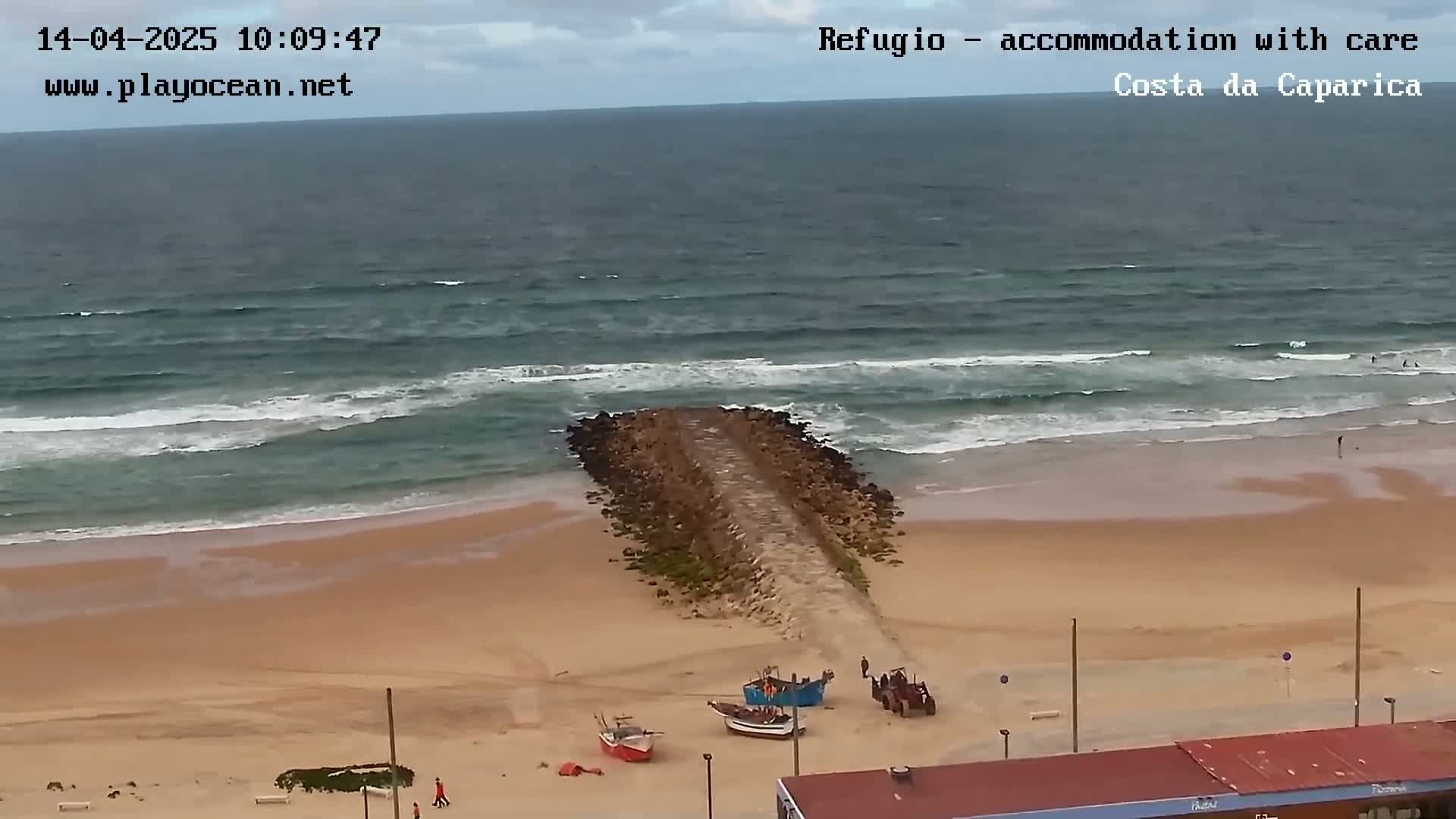 A sandy beach with small waves, several small boats, and a stone jetty extends into the ocean under an overcast sky.