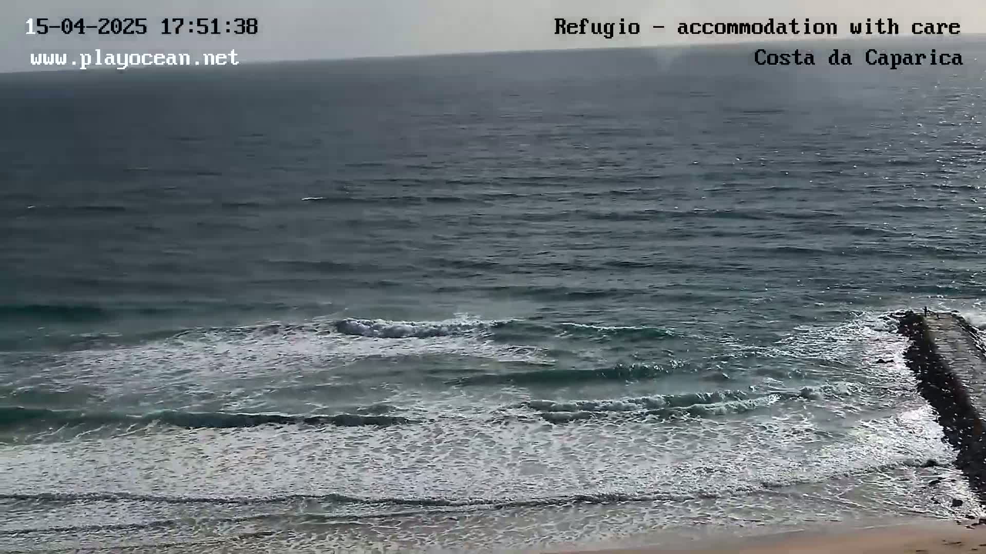 A dark, somewhat overcast day shows moderate waves breaking on a beach near a jetty.