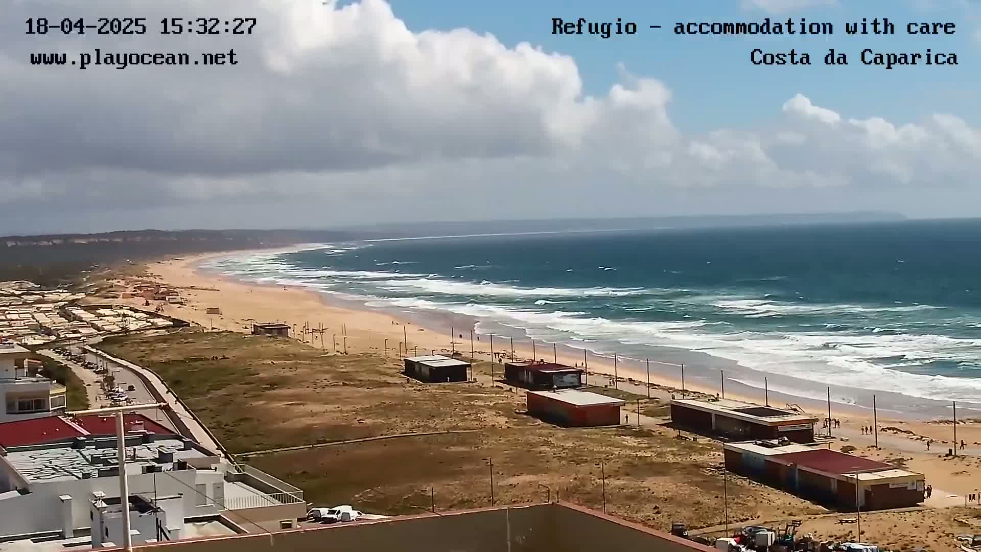 A long sandy beach with moderate waves under a partly cloudy sky is seen from an elevated viewpoint, with buildings and structures visible in the foreground.