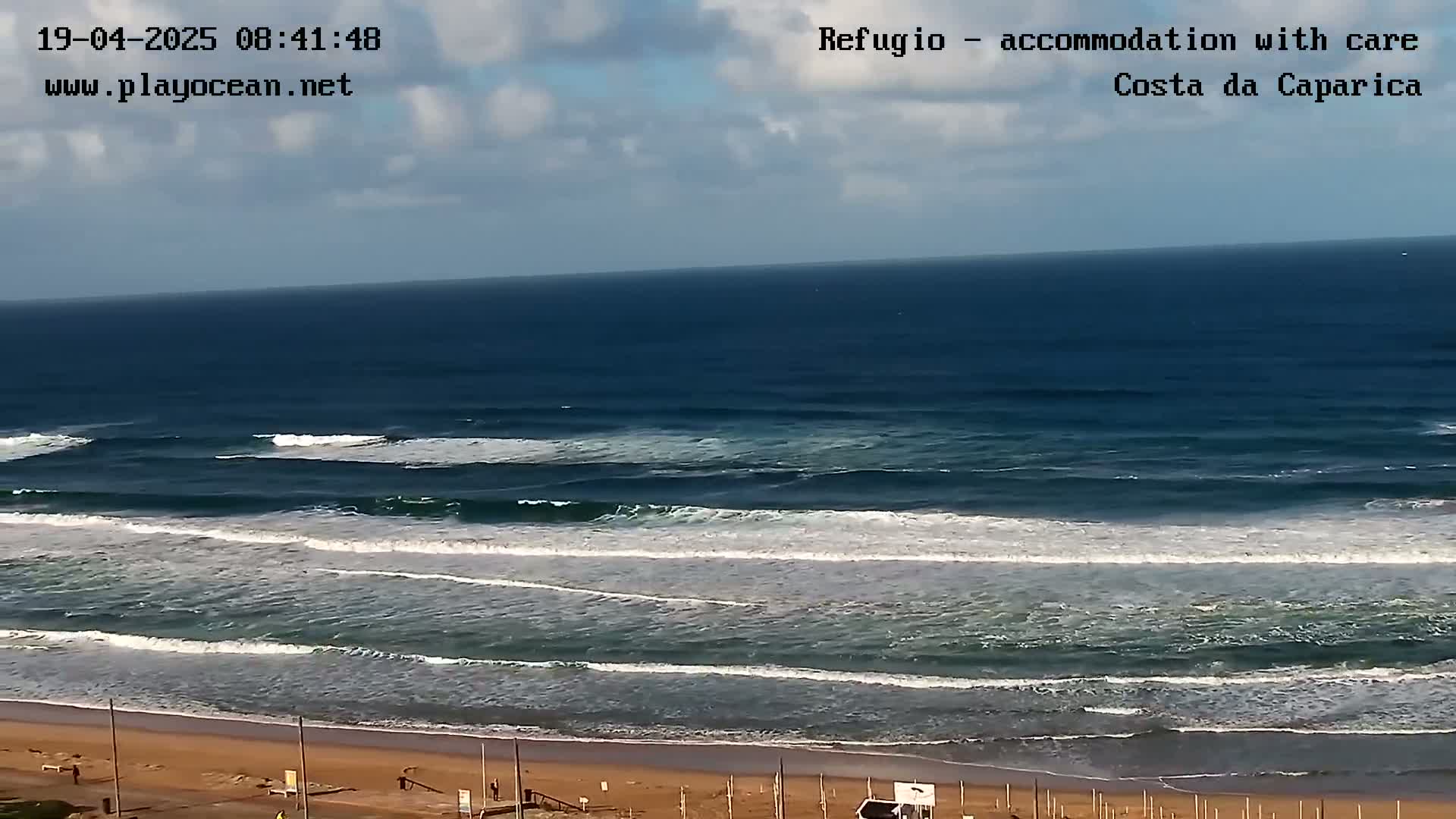 A mostly sunny day shows a wide sandy beach and ocean with gently rolling waves.