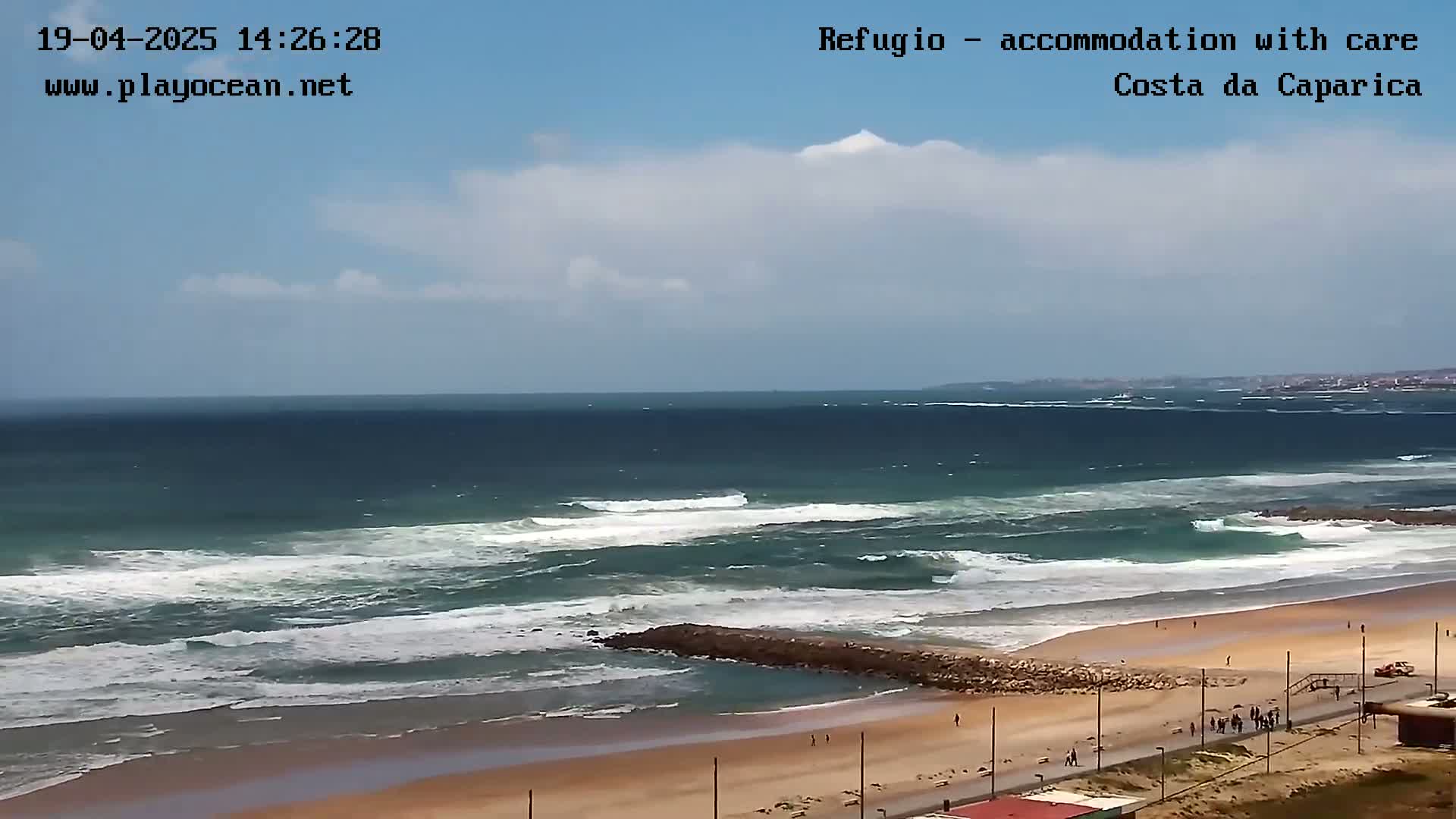 A partly cloudy day shows a wide sandy beach with moderate waves breaking near a rock jetty.