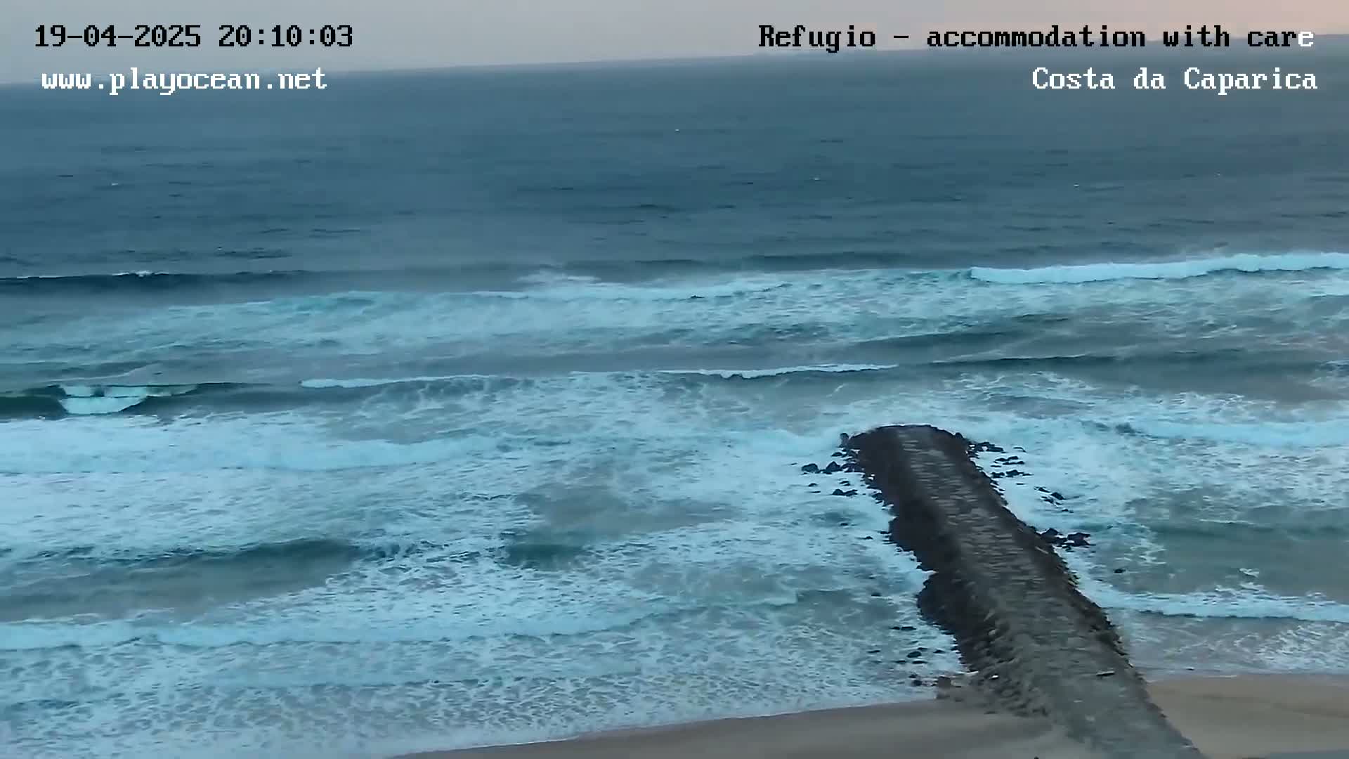 A long, dark jetty extends into a sea with moderate waves under a somewhat overcast sky.