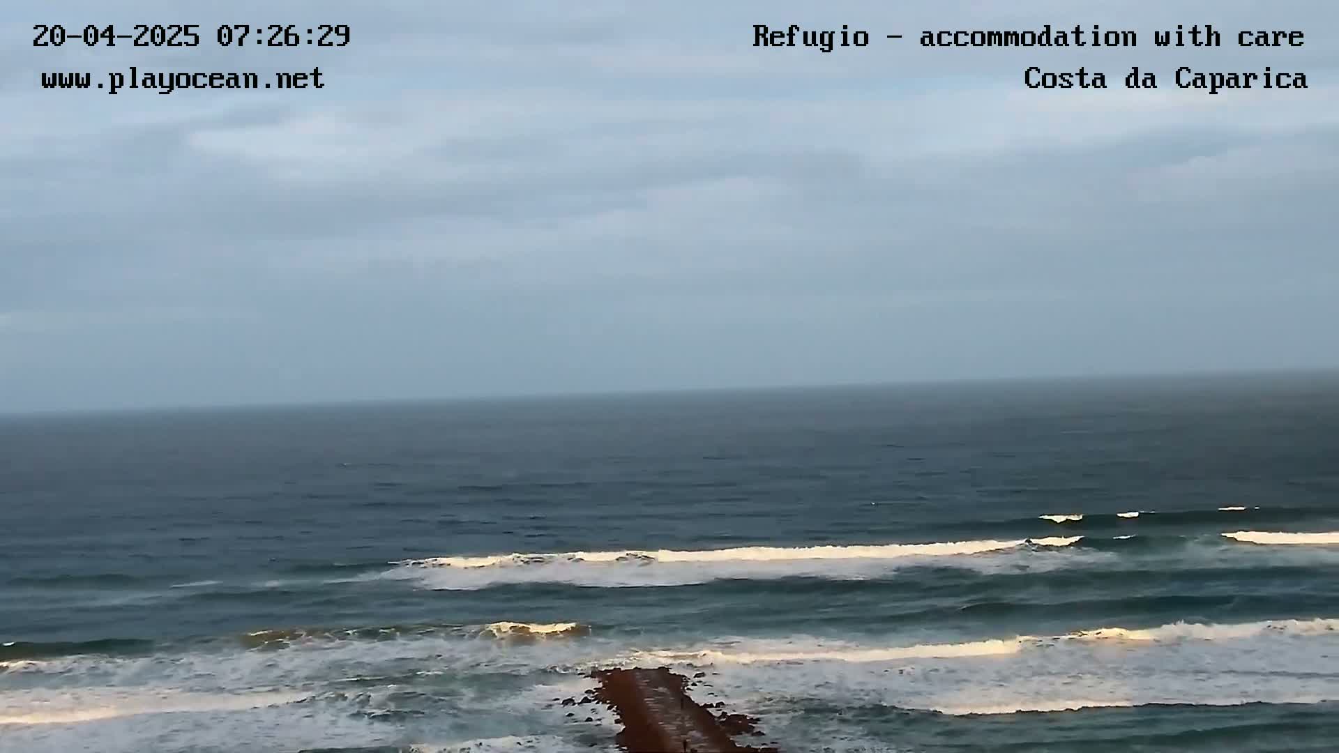 A long, dark-blue ocean with small waves breaking on the shore under an overcast sky is shown, with a long, thin jetty extending into the water.