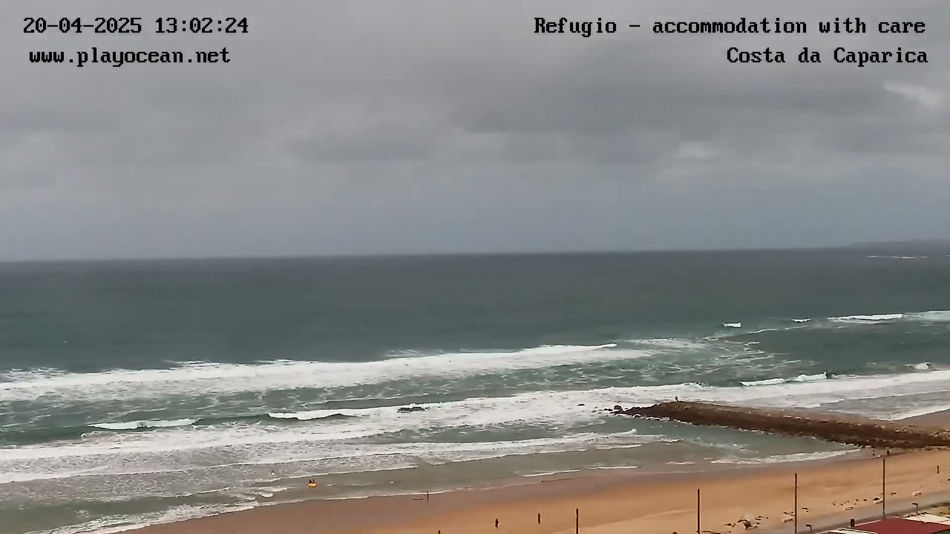 A sandy beach with moderate waves under an overcast sky is seen from an elevated vantage point.