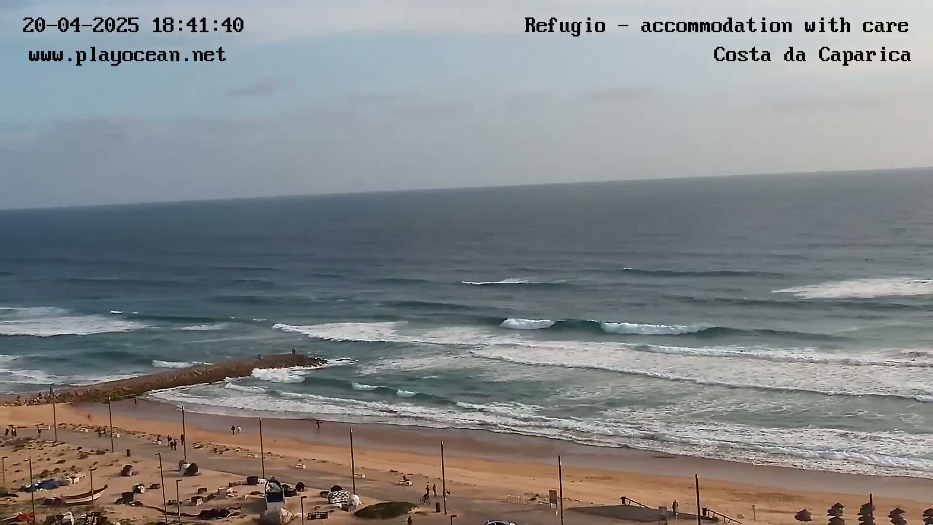 An ocean beach with moderate waves and a mostly clear sky is shown from an elevated perspective.